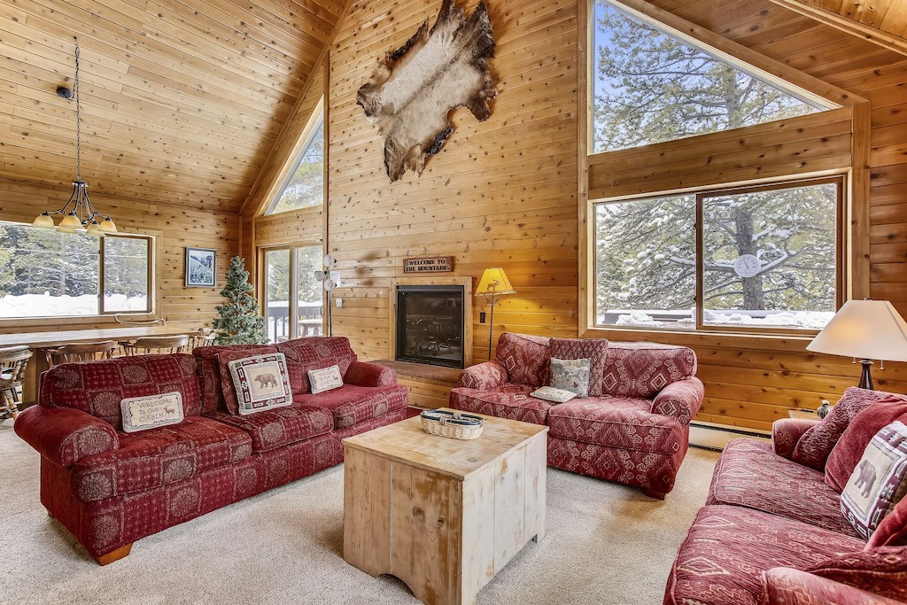Cozy up in your rustic living room with plush sofas, warm fireplace, and stunning forest views through soaring windows.