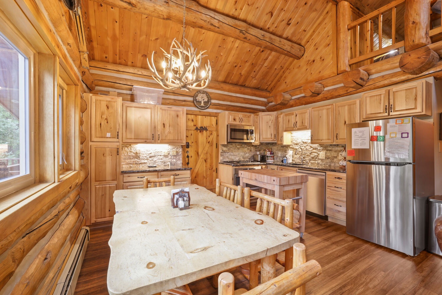 Step into your rustic mountain retreat featuring a fully equipped kitchen with modern appliances, a grand chandelier overhead, and handcrafted log furniture perfect for gathering around home-cooked meals.