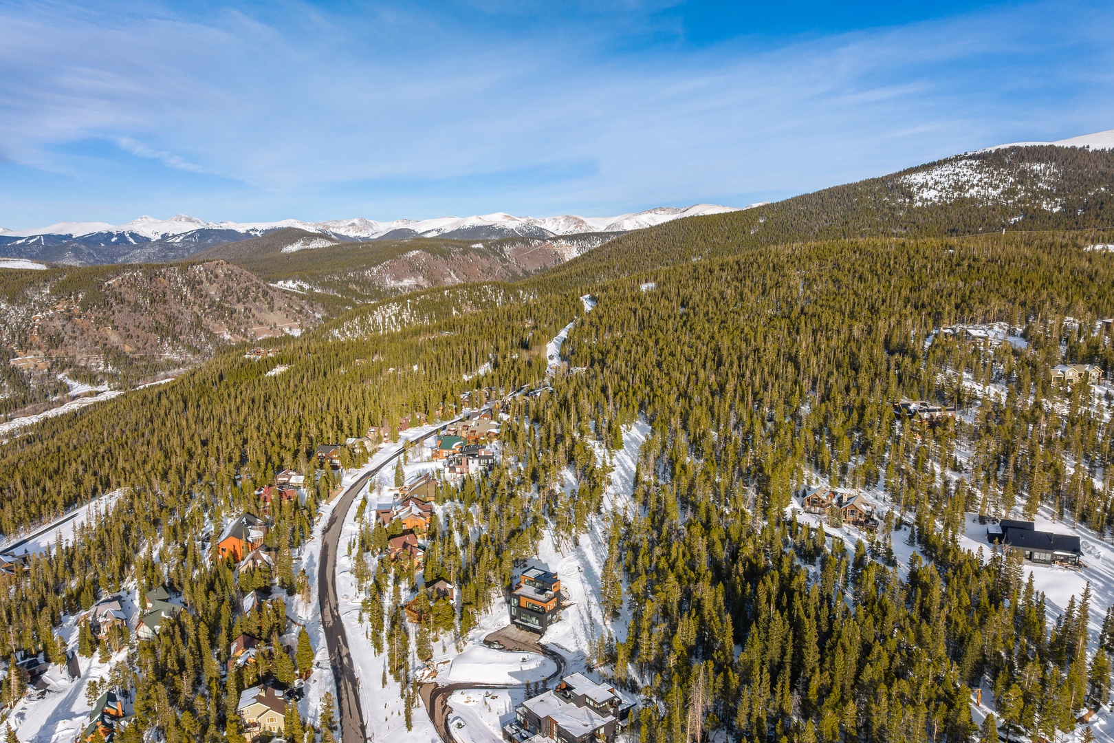 Aerial view of a mountain resort community nestled among snow-covered evergreen forests with spectacular Rocky Mountain peaks in the distance.