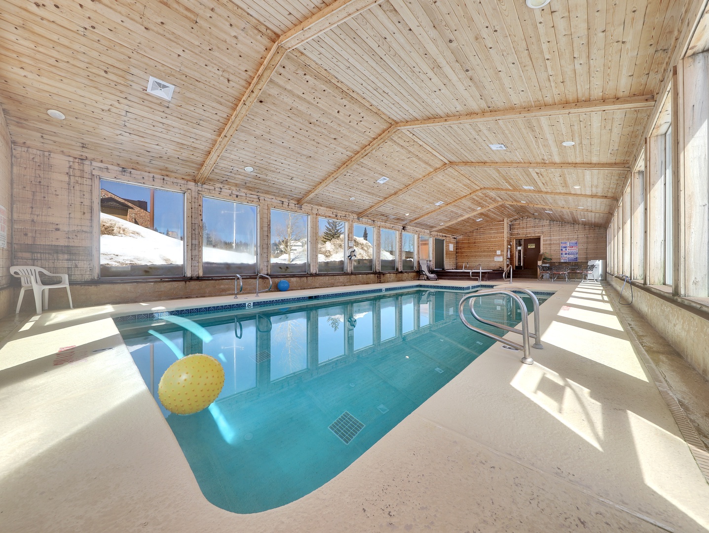 Indoor swimming pool enclosed within natural wood beams, featuring expansive windows framing snow-covered mountain views and bright sunlight—ideal for year-round relaxation.
