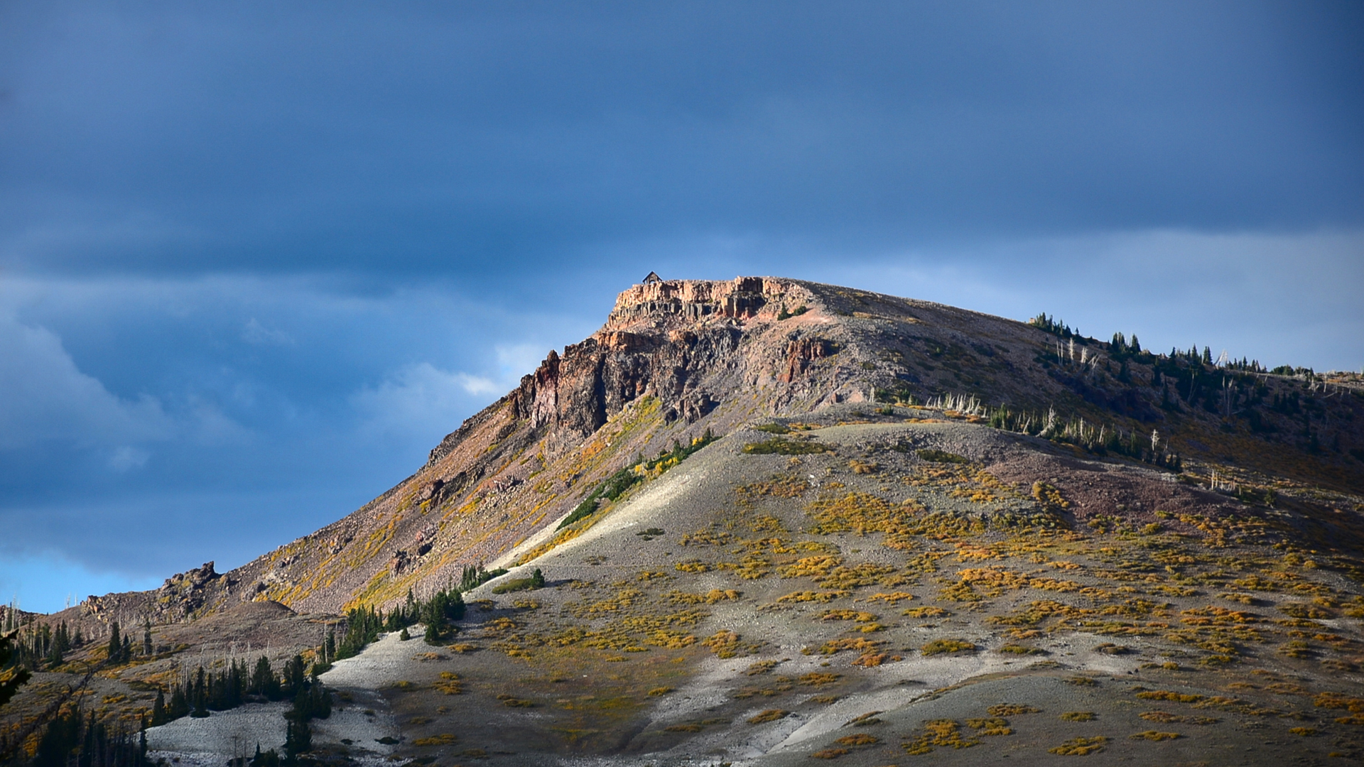 A dramatic mountain peak rises above the surrounding landscape, with rugged rock formations catching golden light and scattered evergreens dotting the slopes.
