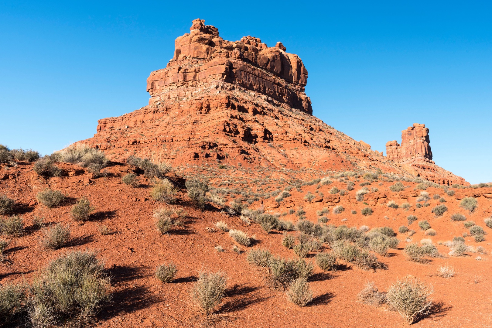 A dramatic red rock formation rises against clear blue skies, surrounded by desert vegetation and offering striking natural scenery in the surrounding area.