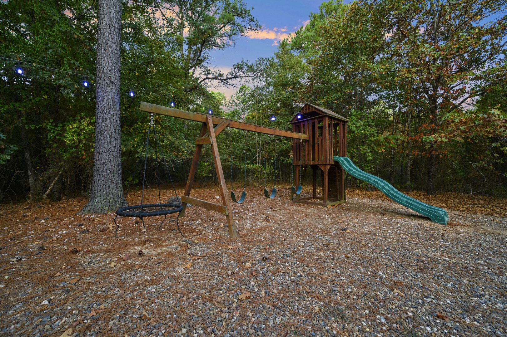 This playground nestled in the woods features swings, a slide, and a climbing structure, perfect for creating family memories under string lights and open sky.