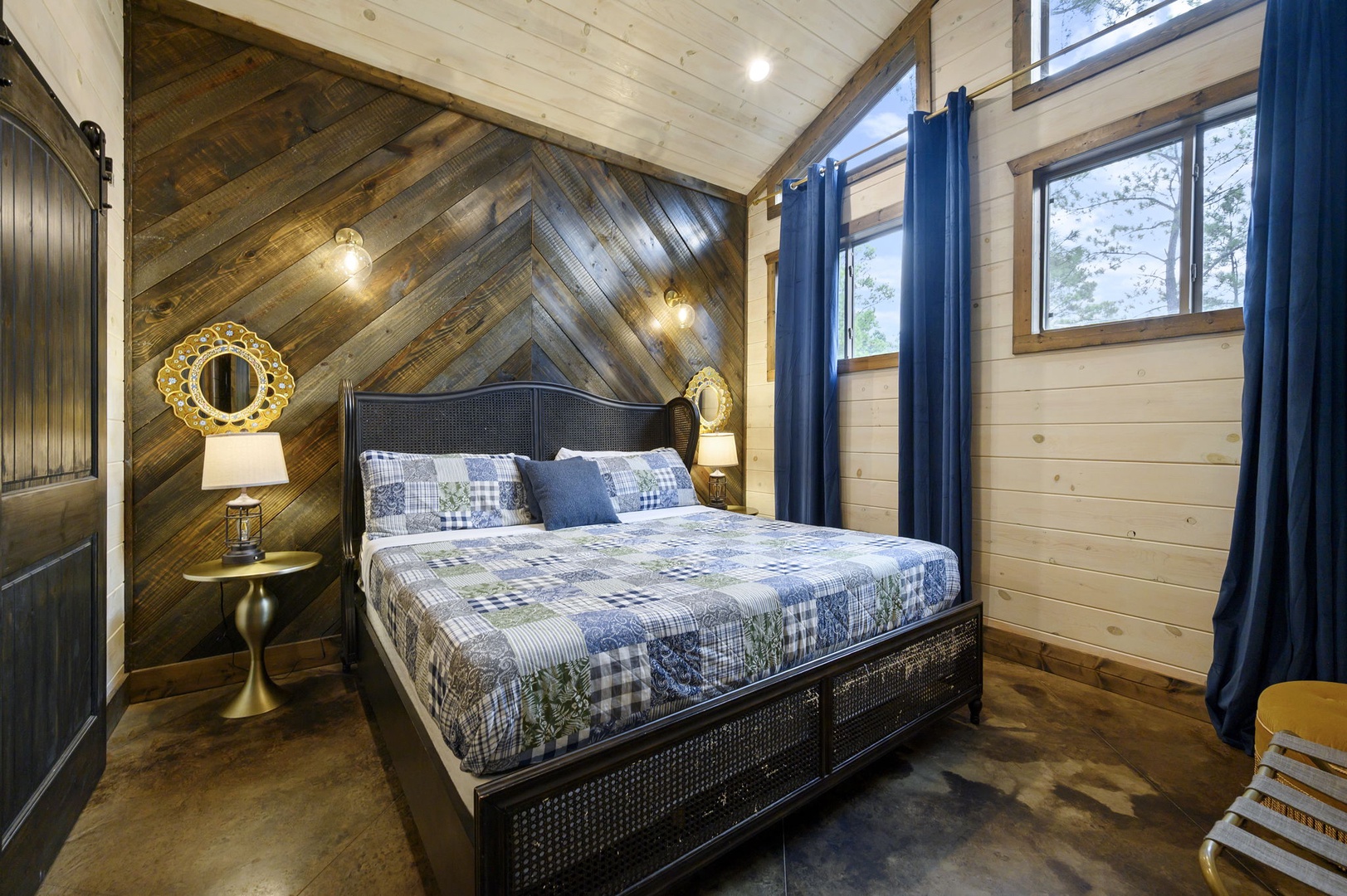 Your cozy bedroom features a handsome accent wall of chevron-patterned wood, a plush bed dressed in blue and white, and natural light streaming in through the windows. Warm lighting and thoughtful decor create a restful retreat.