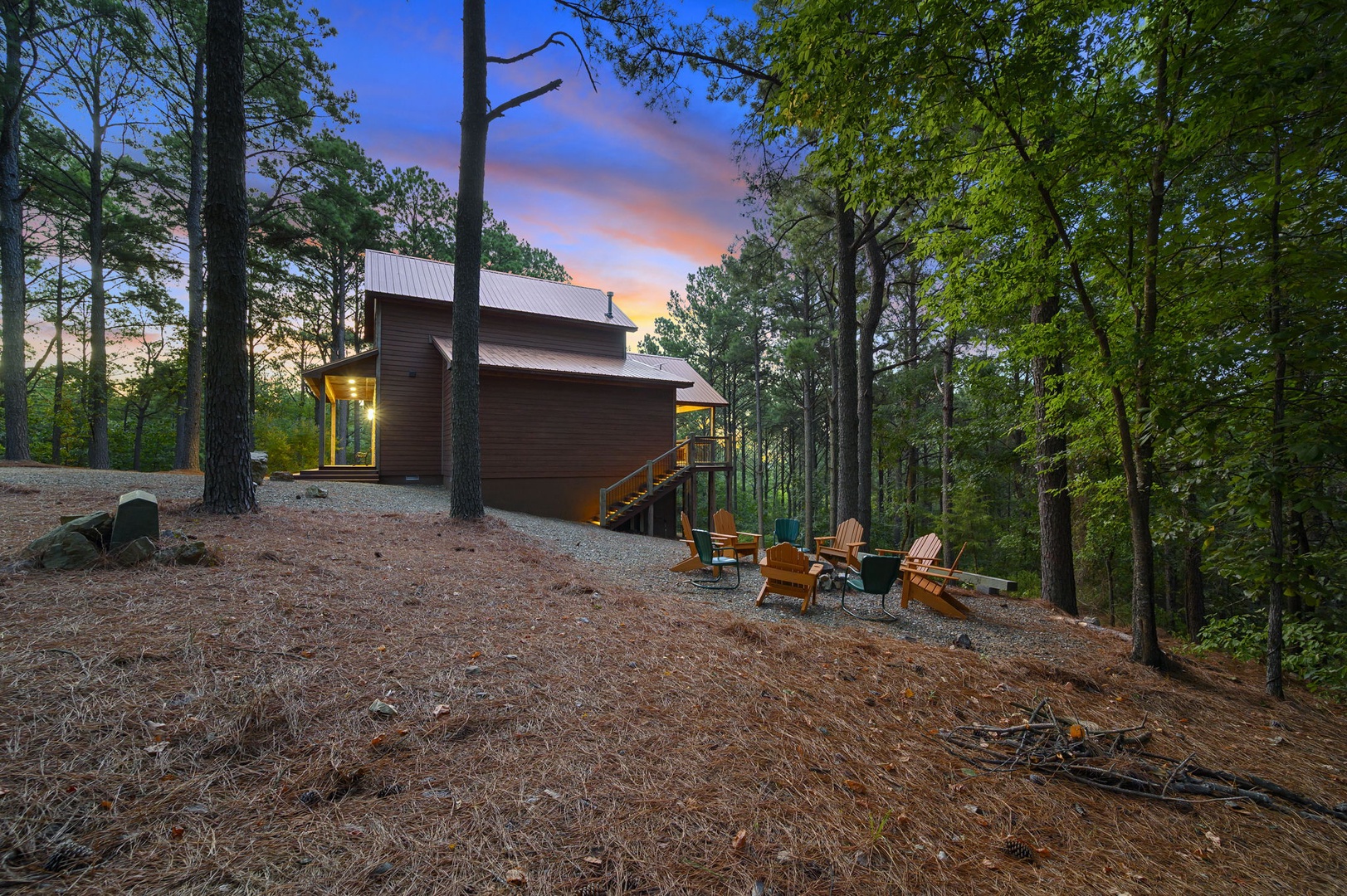 Modern cabin nestled in towering pines with a vibrant sunset backdrop, surrounded by lush forest. A welcoming fire pit and colorful Adirondack chairs invite peaceful evenings under the trees.