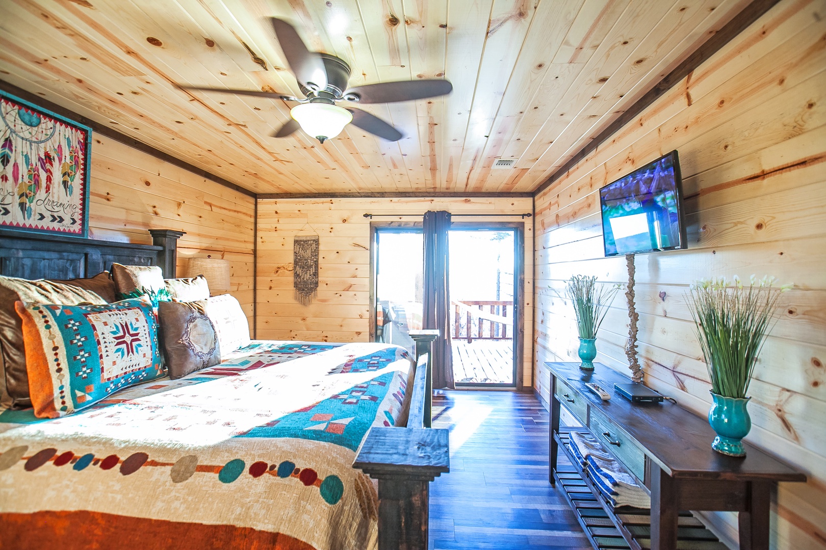 Relax in your cozy cabin bedroom with rustic wood walls, vibrant southwestern decor, and peaceful balcony access for mountain air.