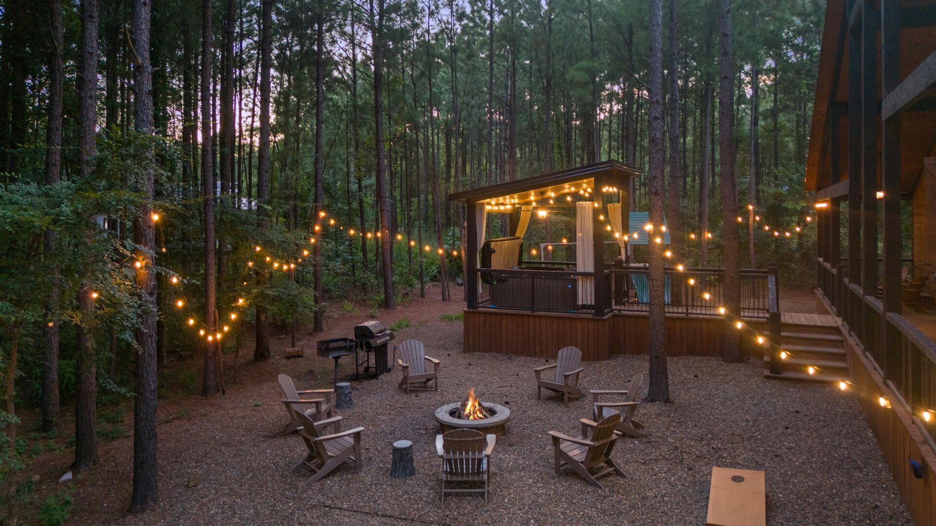 The wooded property features an outdoor entertaining area with string lighting, fire pit seating, covered bar pavilion, and multiple outdoor structures surrounded by tall pines at dusk.