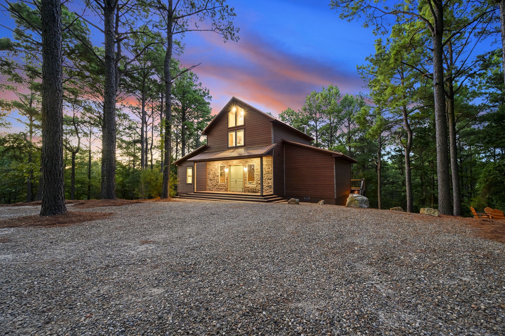 A modern woodland retreat featuring stone accents and warm lighting, set among tall pines with ample gravel parking—photographed at twilight as dusk paints the sky in soft pastels.