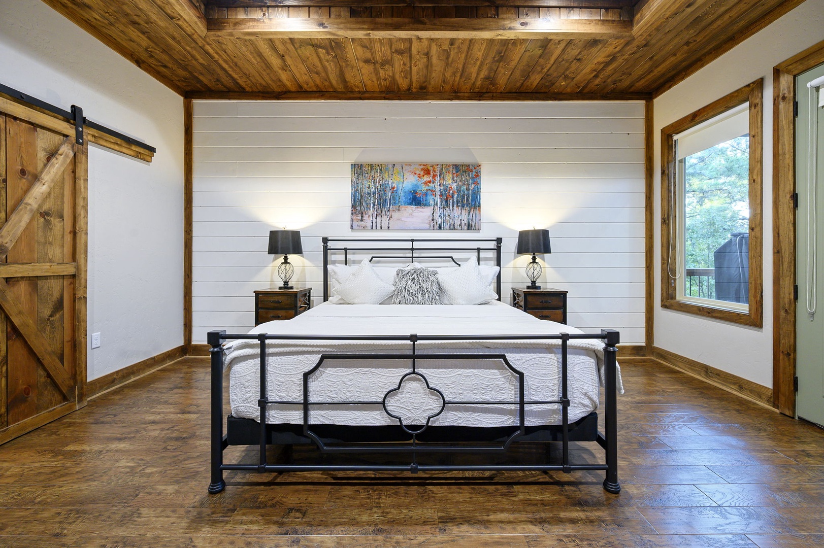 Rest easy in your spacious bedroom with rustic wood-beam ceilings, a charming barn door, and a cozy king bed framed by warm natural light streaming through the window.