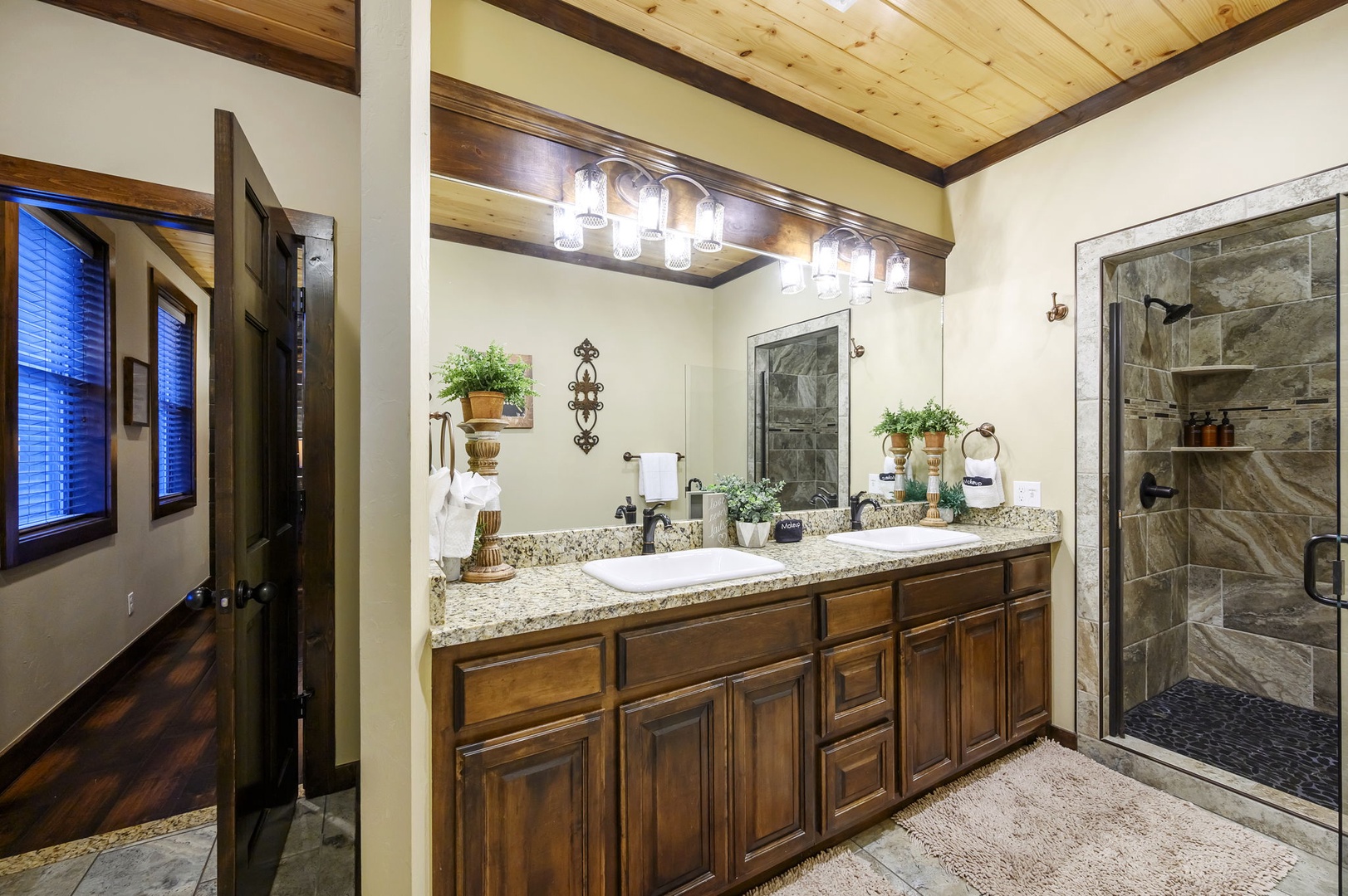 Freshen up in your spacious bathroom, featuring dual vanities with granite countertops, a stylish stone-tiled walk-in shower, and warm wood accents under a stunning timber-beamed ceiling.