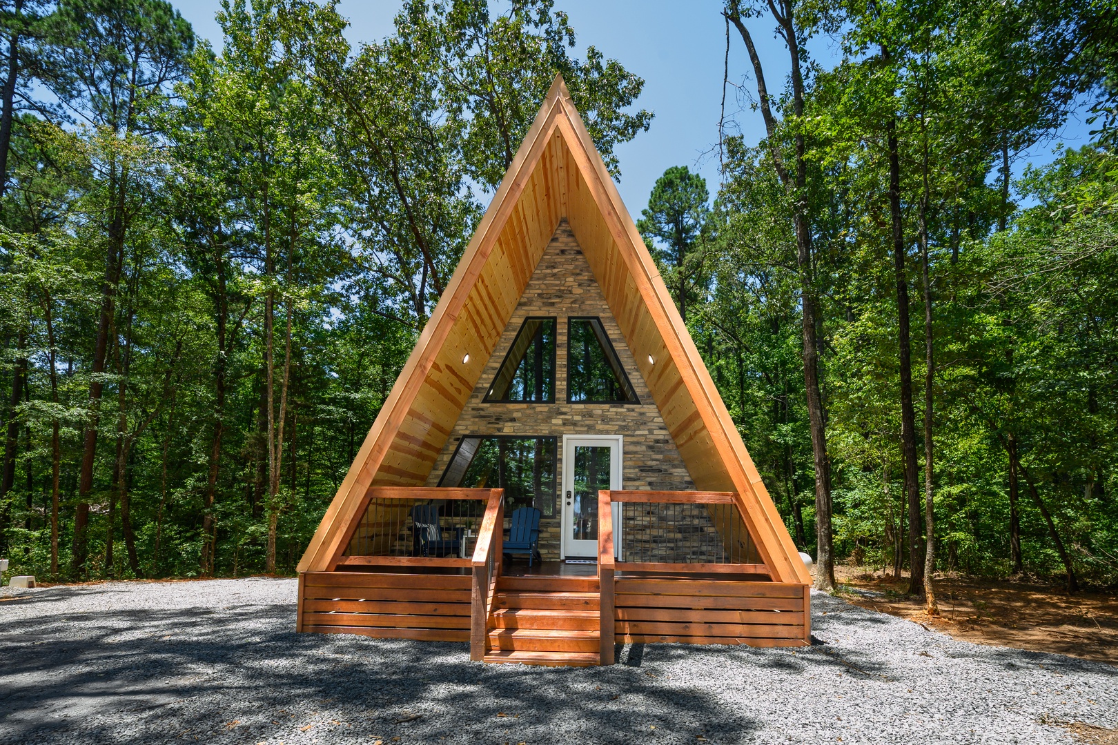 A striking A-frame cabin nestled in the woods, featuring warm wood tones and large windows that invite natural light. The welcoming front deck with wooden stairs provides an ideal spot for morning coffee surrounded by towering trees.