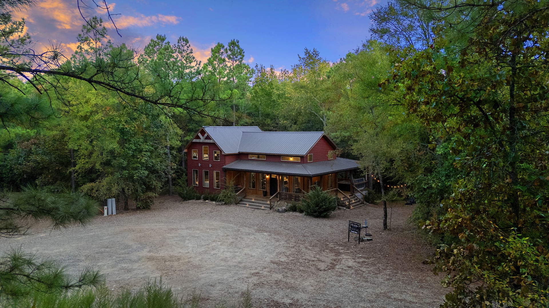 A charming red barn-style property nestled among lush trees at dusk, offering a peaceful wooded retreat with warm lighting and rustic appeal.