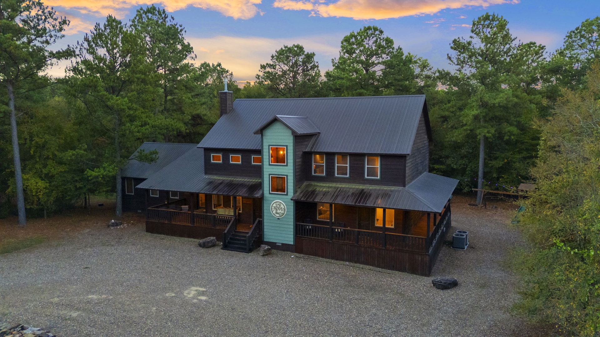 The property stands nestled among towering pines as evening light warms the windows, with generous parking and a welcoming arrival experience.
