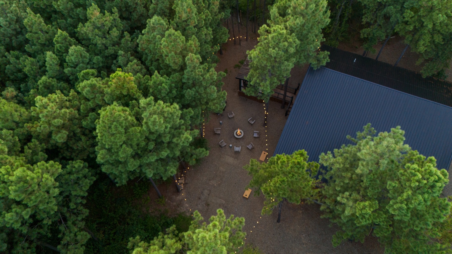 A secluded retreat nestled deep in lush forest, where paths wind through towering trees to gathering spaces with fire pit, seating, and shaded dining area.