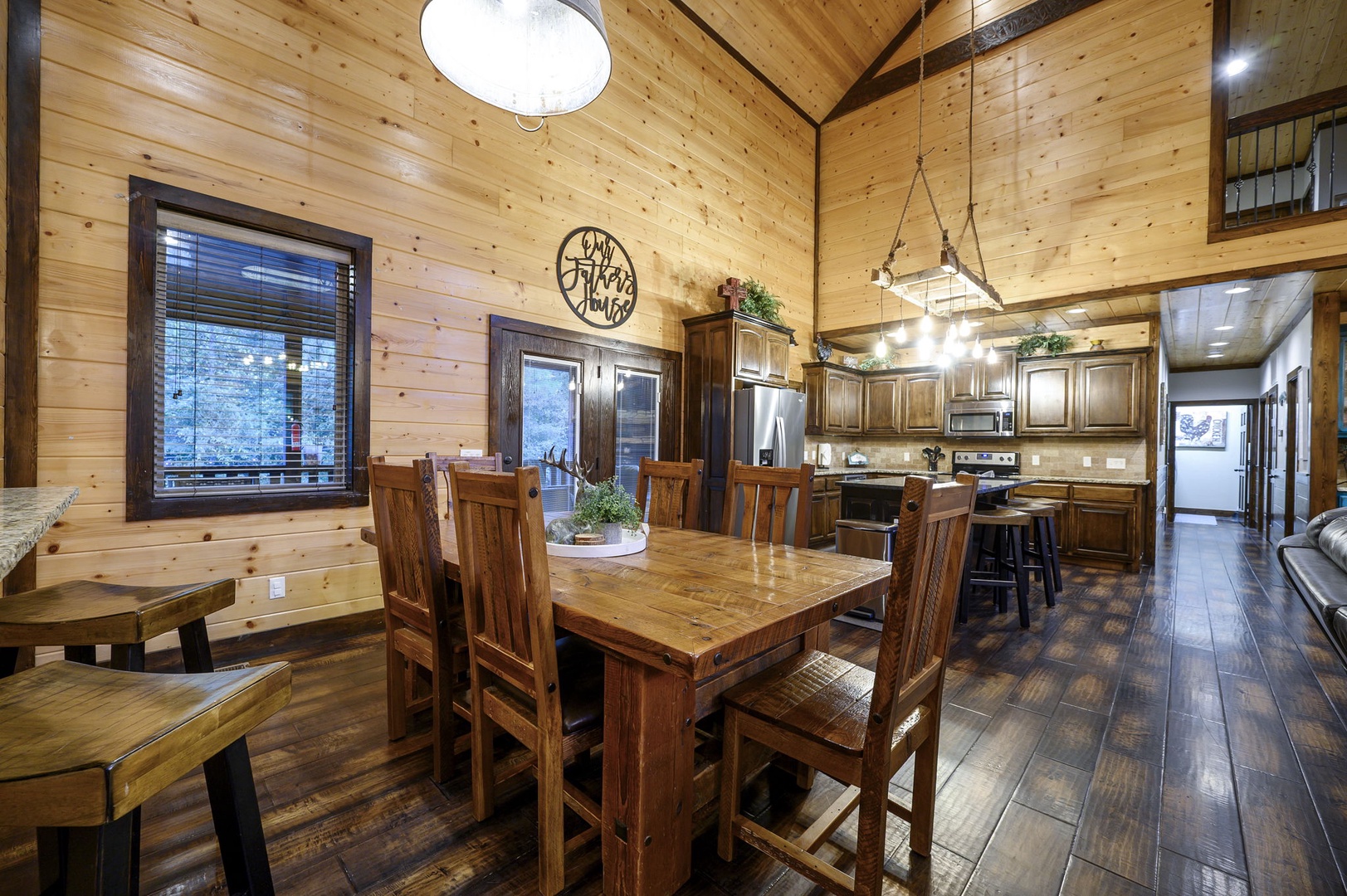 Your mountain retreat opens into a warm, vaulted great room where rustic wood beams soar above a spacious dining table and fully-equipped kitchen, bathed in natural light from large windows.