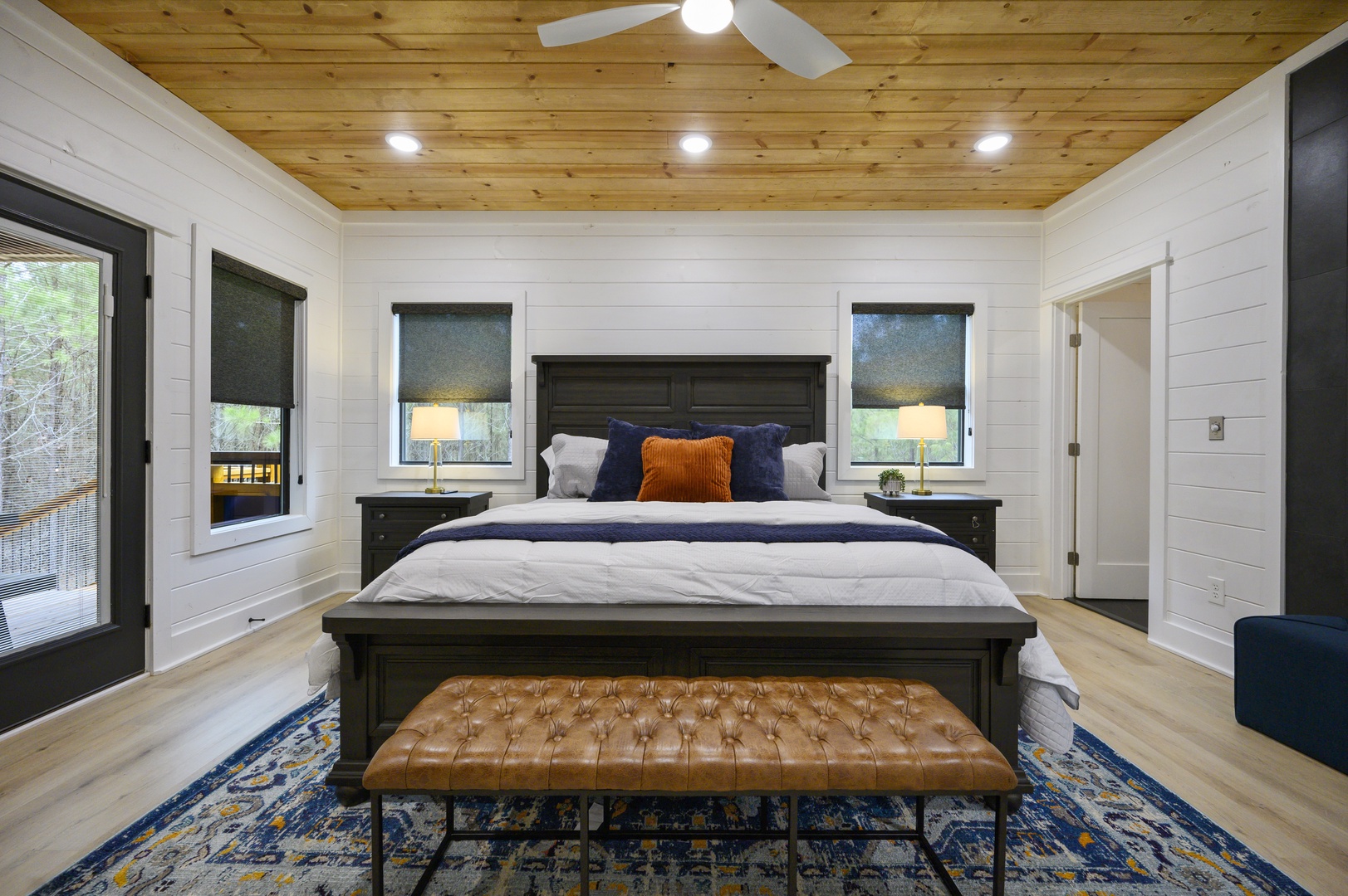 Unwind in this stunning bedroom with rustic wood ceiling, cozy bedside lamps, and peaceful forest views through every window.