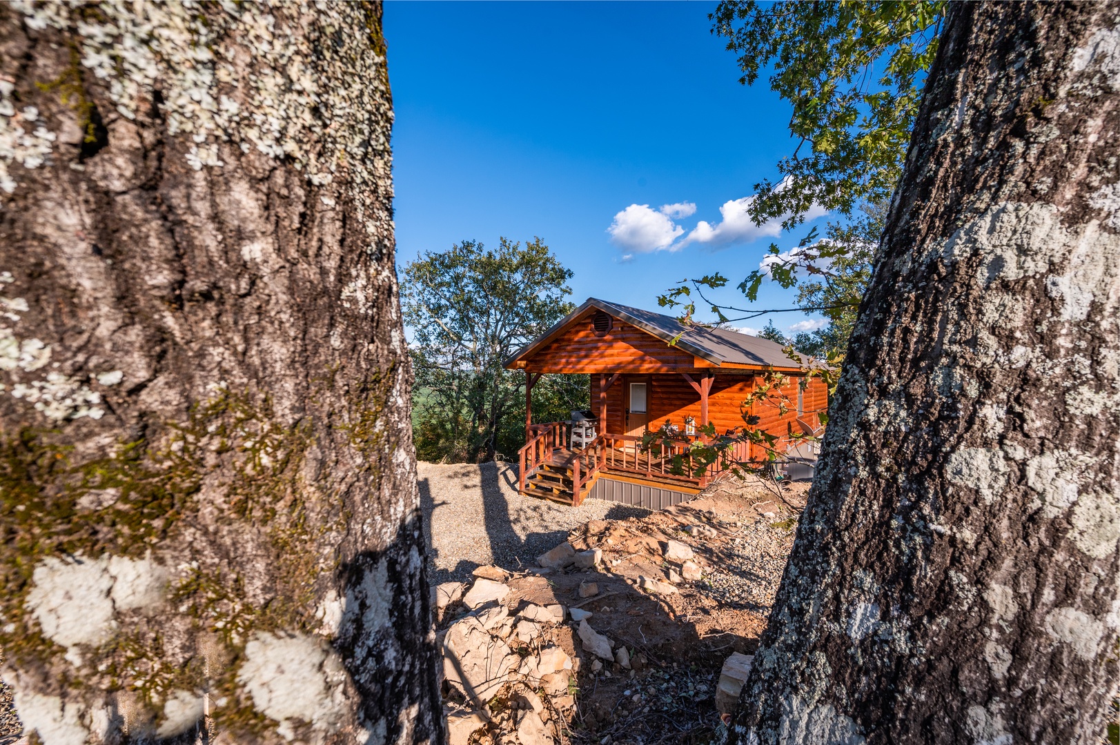 Rustic wooden cabin nestled among towering trees, featuring a welcoming covered deck surrounded by natural stone formations.