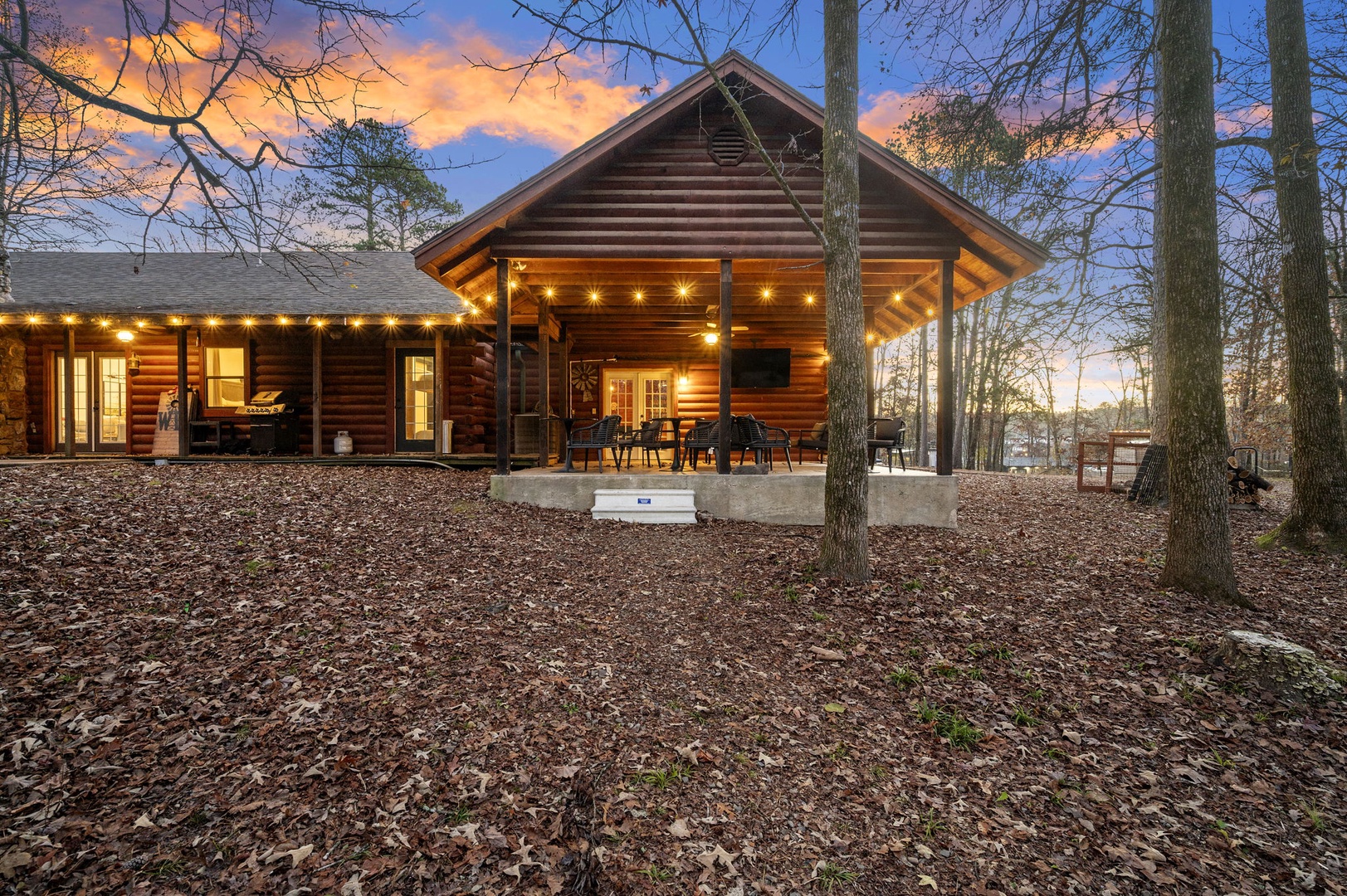 Beautiful log cabin nestled among towering trees, featuring a welcoming covered porch with string lights creating perfect ambiance for evening relaxation.