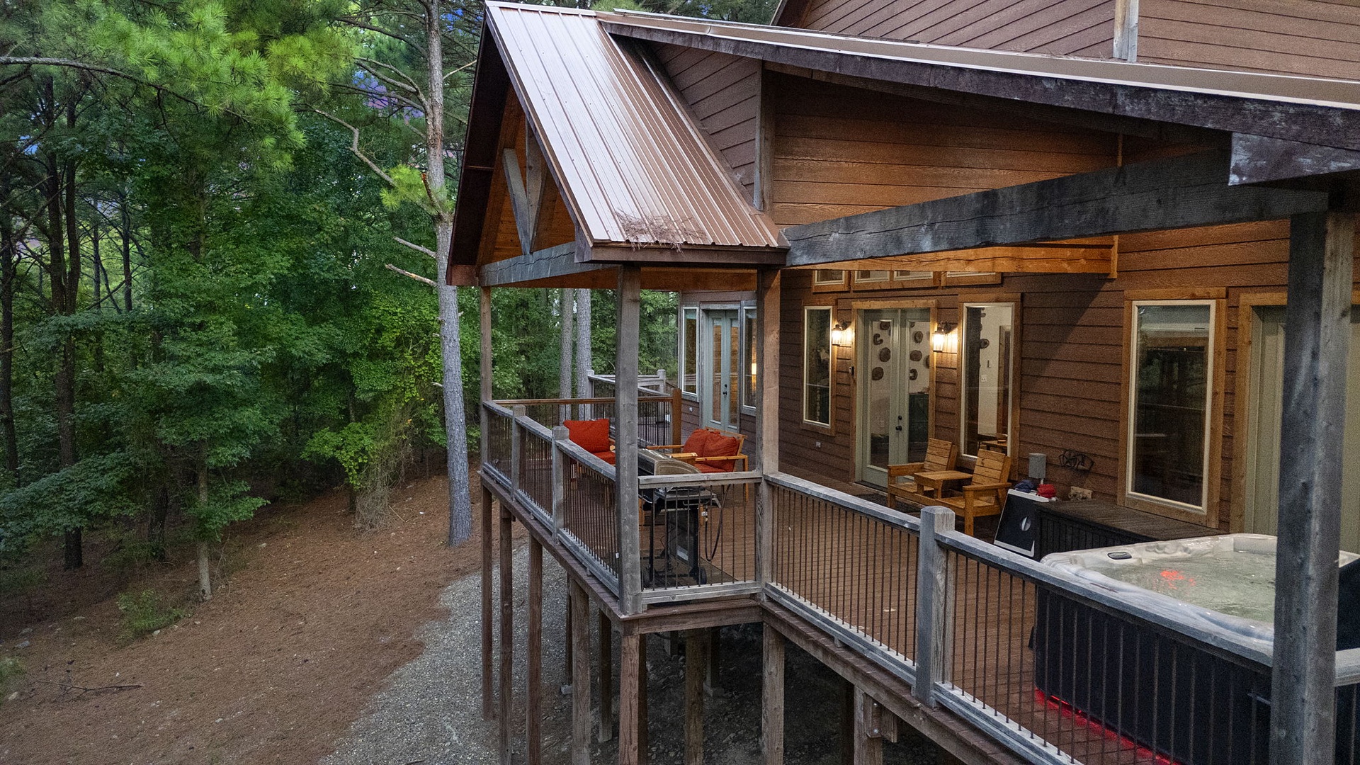 A contemporary cabin exterior nestled among towering pines, featuring a spacious deck with a hot tub and relaxing seating, offering peaceful forest views.