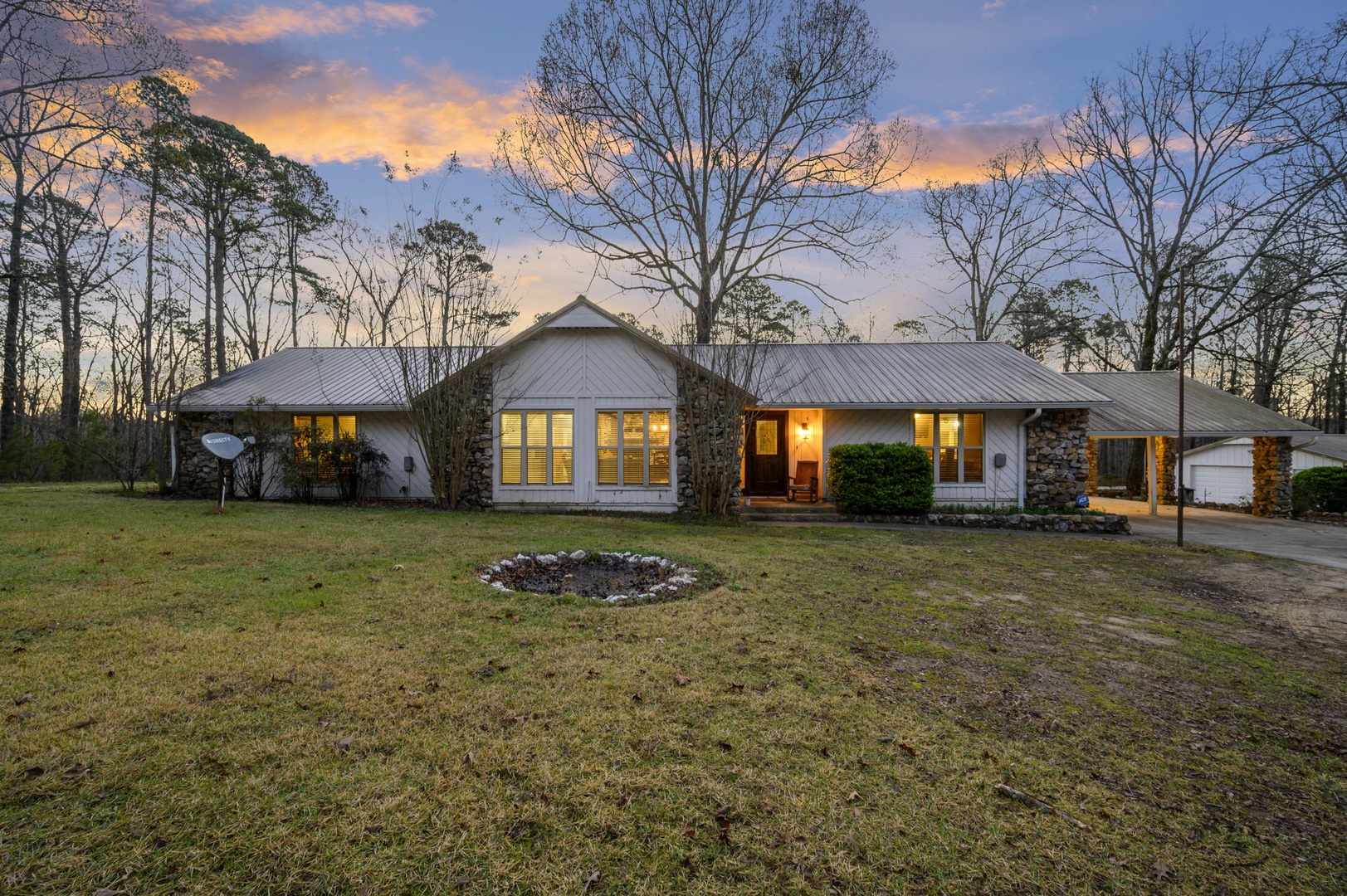 Modern ranch-style home nestled among towering trees with stone accents and metal roofing, photographed during golden hour.
