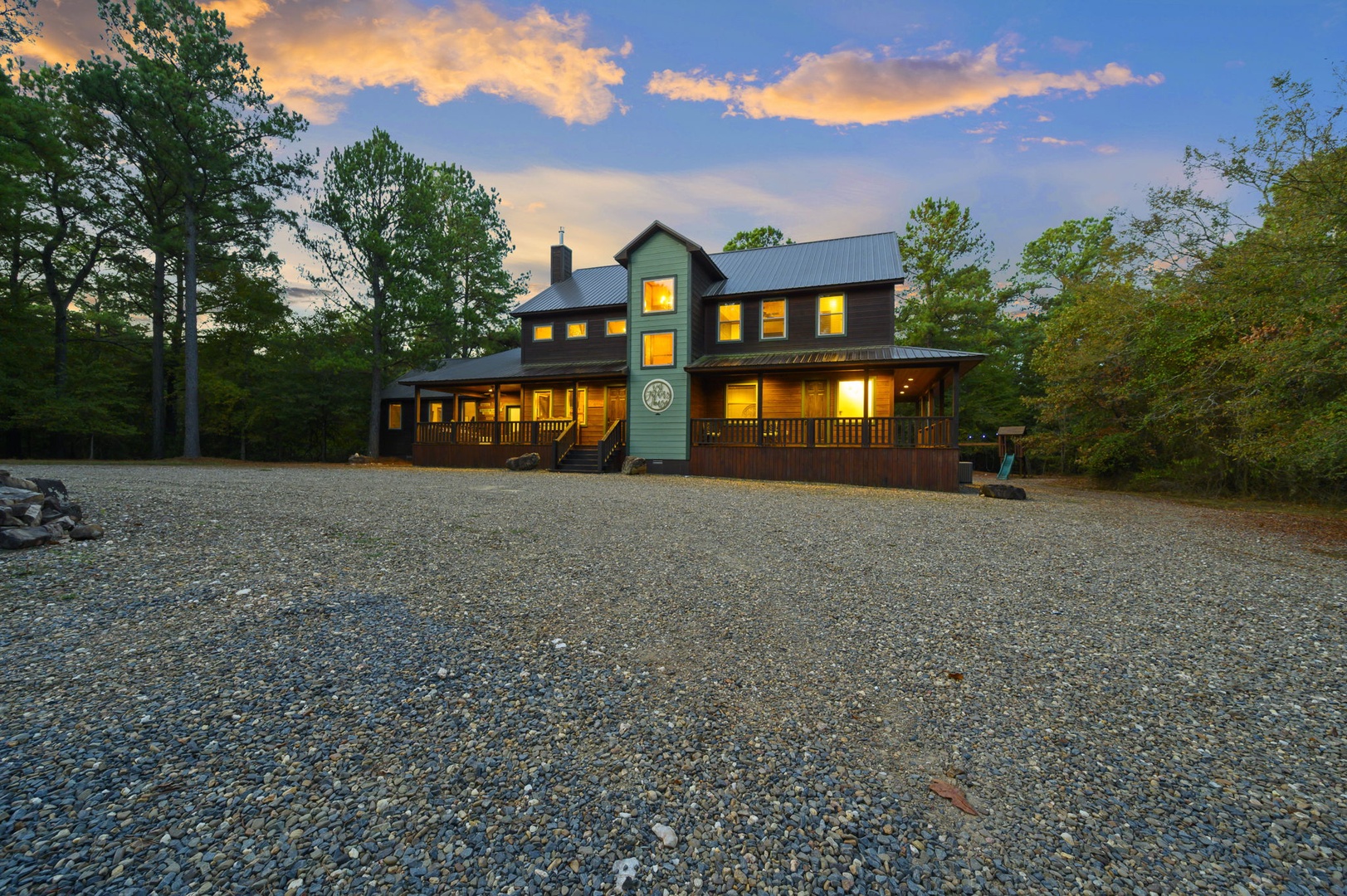 A spacious modern farmhouse-style retreat nestled in a peaceful wooded setting, featuring generous parking and welcoming covered porches, captured during a beautiful sunset.