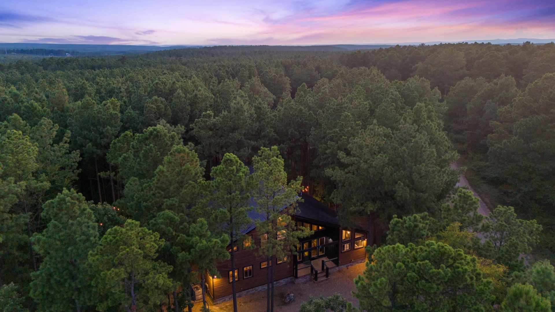 A stunning forest cabin property stands nestled among towering trees at twilight, surrounded by pristine woodland as far as the eye can see.