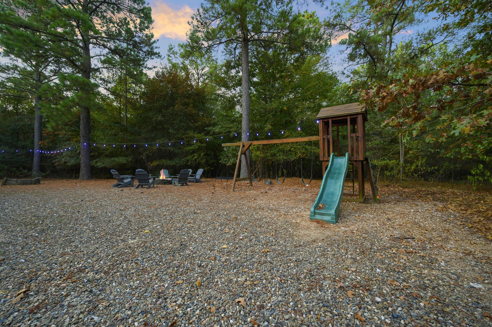 Spacious backyard retreat surrounded by tall trees features a playground, swings, fire pit seating, and twinkling string lights for peaceful outdoor evenings.
