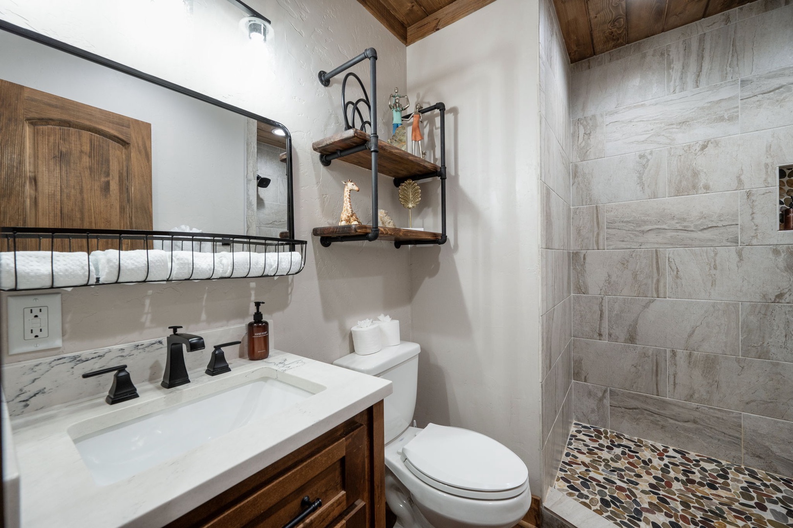 Step into your beautifully designed bathroom featuring a walk-in shower with rustic stone tile, warm wooden accents, and thoughtful touches like industrial shelving and soft rolled towels waiting for you.