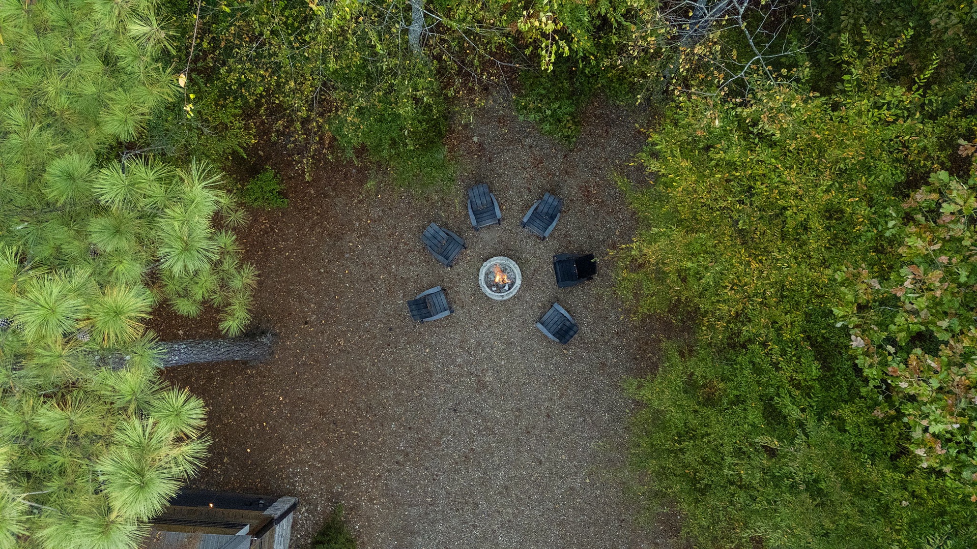 An aerial view of the fire pit area, featuring a cozy circle of Adirondack chairs arranged around a central fireplace, nestled within a peaceful wooded setting.