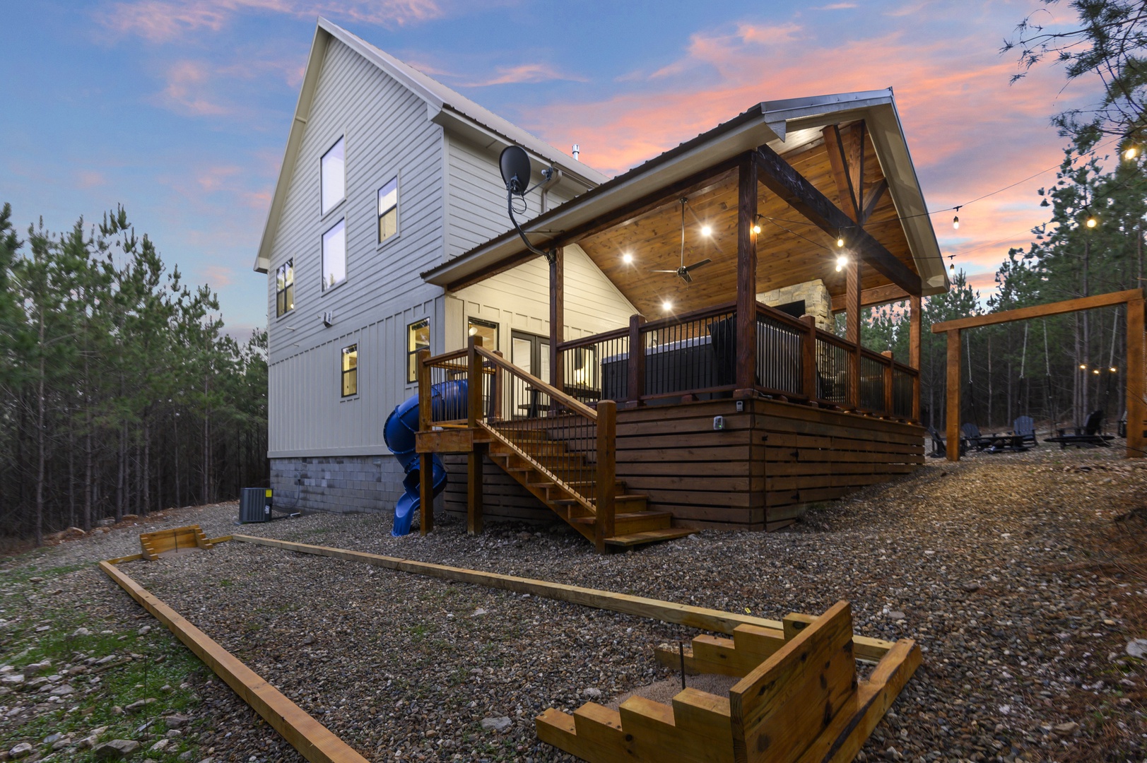 A stunning modern retreat featuring a spacious deck with outdoor lighting and playground, surrounded by peaceful wooded landscape at dusk.