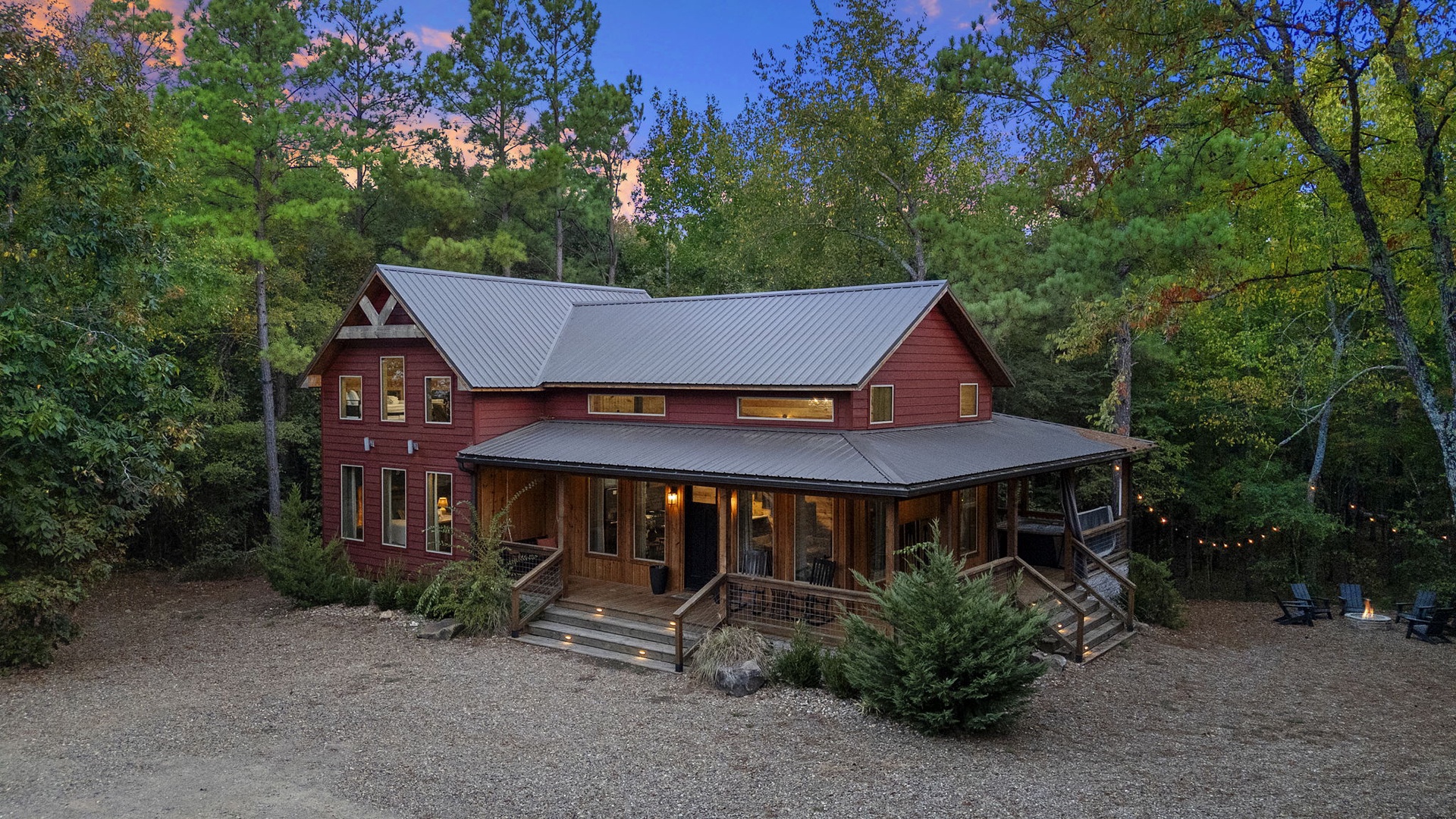 A welcoming red farmhouse-style retreat surrounded by towering pines and deciduous trees in a peaceful forest setting.