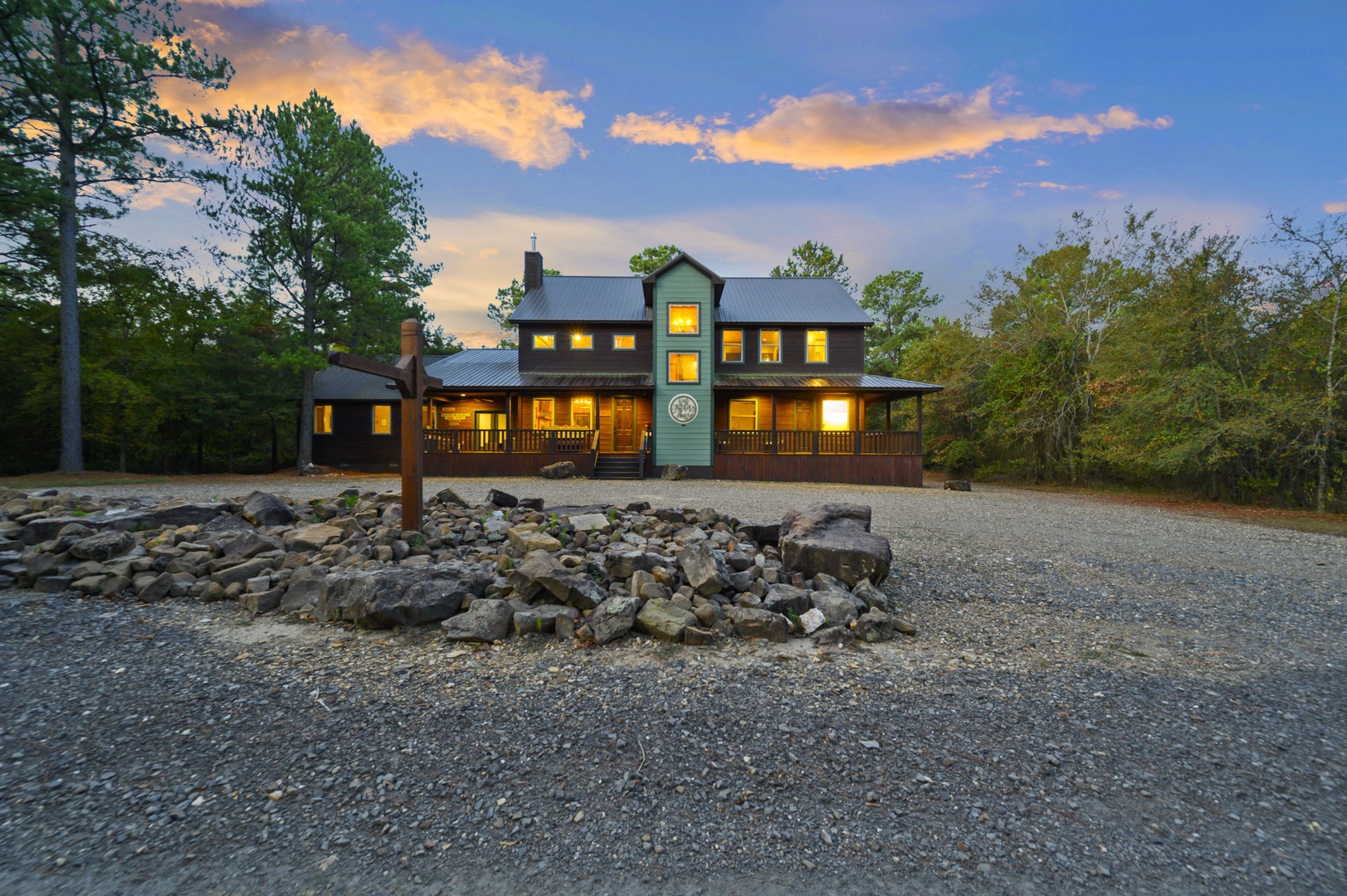 Spacious mountain farmhouse-style retreat nestled among tall pines and natural wooded surroundings, featuring a decorative rock fire pit and ample parking on arrival.