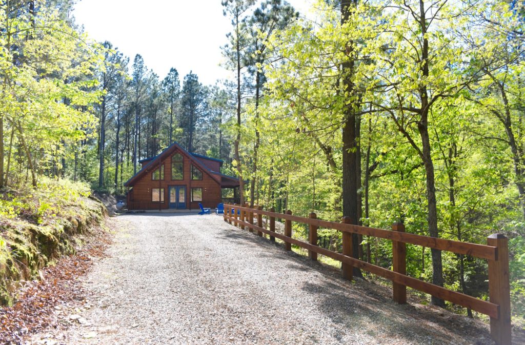 A rustic log cabin nestled among towering trees marks the entrance to this peaceful retreat. The gravel driveway and wooden fence guide visitors to their woodland getaway.