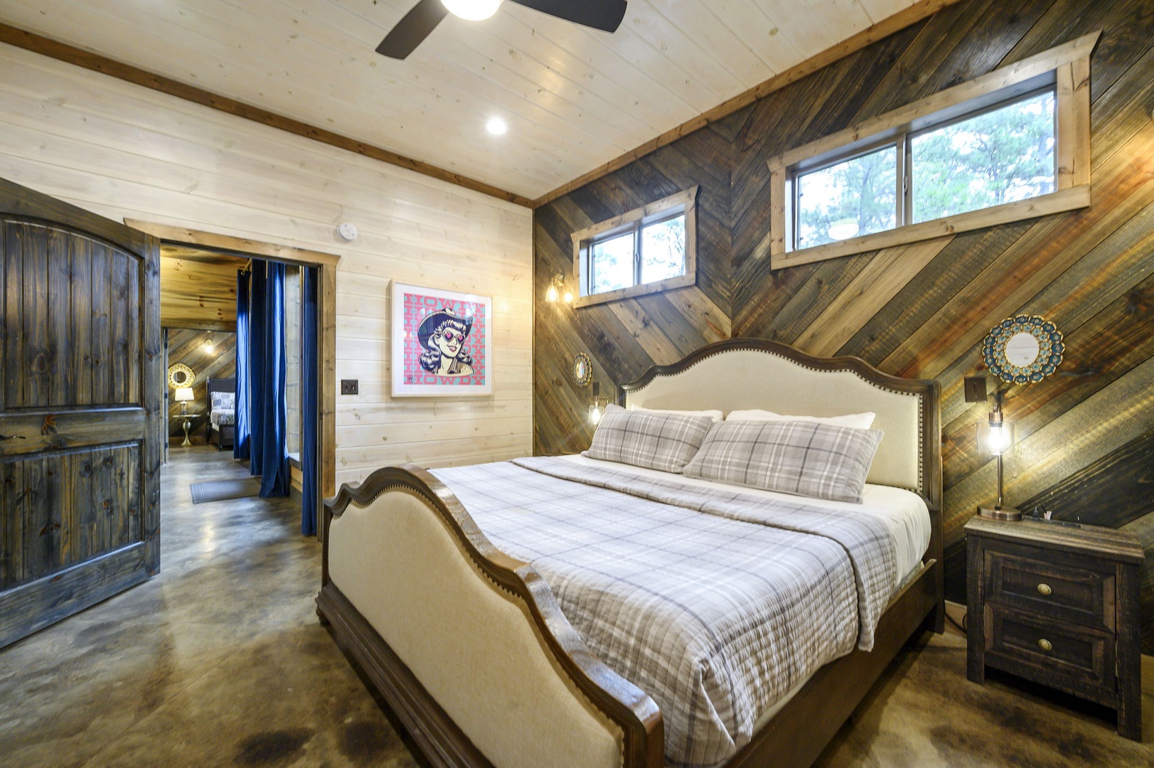 Sink into this inviting bed with its elegant upholstered headboard and cozy plaid linens. The striking diagonal wood accent wall and ceiling fan add rustic charm, while natural light streams through the windows to wake you gently each morning.