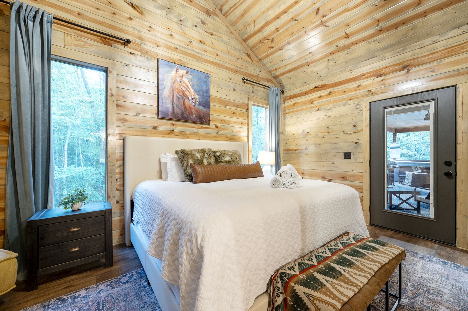 Your cozy cabin bedroom combines rustic wood plank walls and vaulted ceilings with a plush bed ready for restful nights. Step through the glass door to your private outdoor escape.