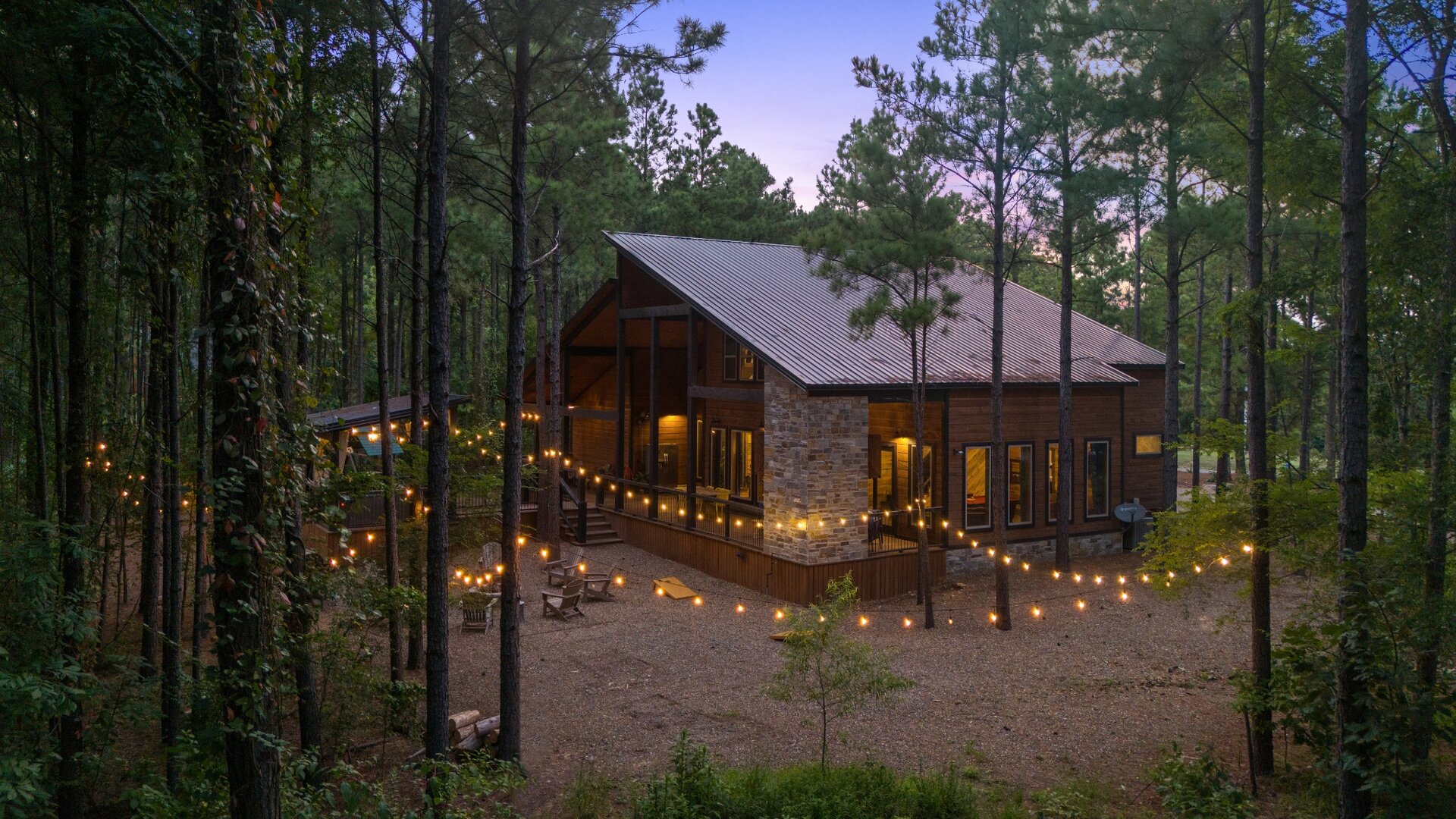 A welcoming cabin nestled among towering pines at dusk, with string lights glowing warmly around the spacious deck and entrance.