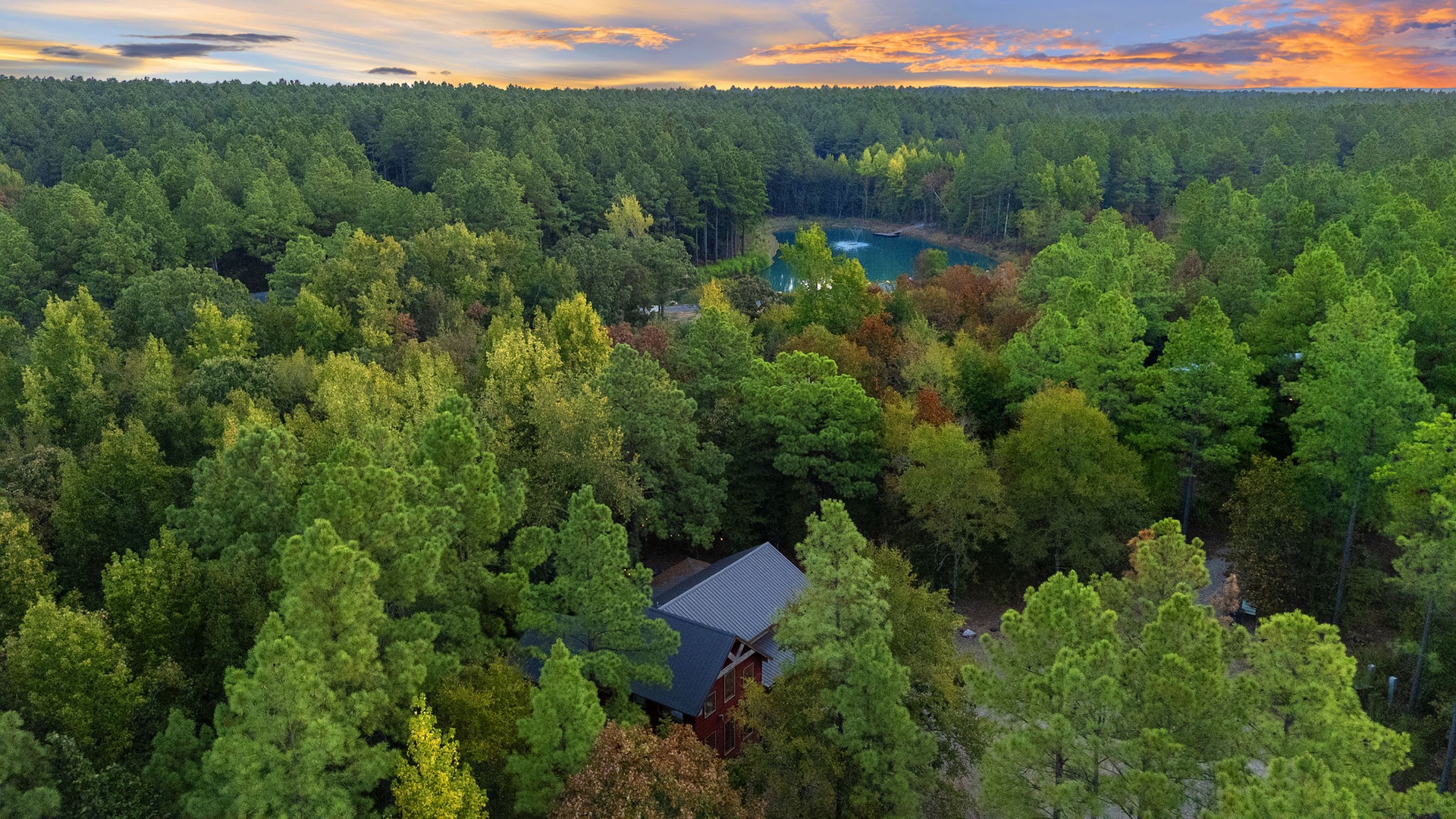 An aerial view captures the property nestled within dense forest, with a tranquil lake visible through the trees and vibrant sunset skies overhead.