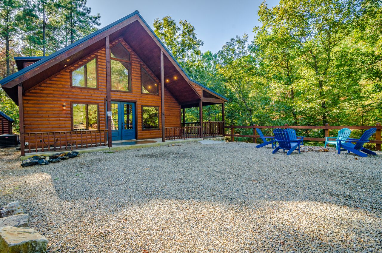 A handsome log cabin featuring a bright blue door welcomes arrivals, surrounded by towering trees and spacious gravel parking. The A-frame design and cheerful Adirondack chairs add rustic charm.