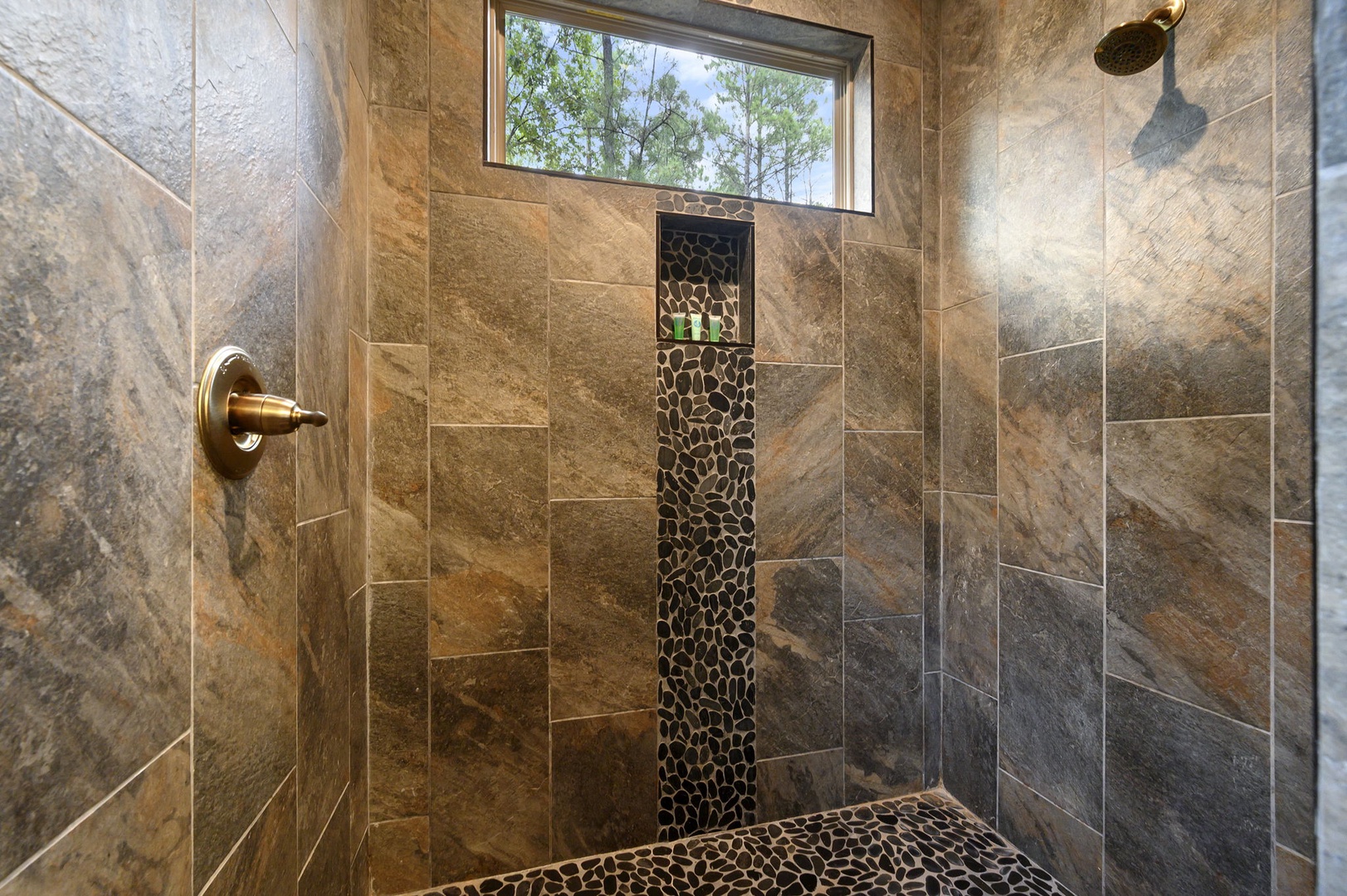 Step into your beautifully tiled walk-in shower with elegant bronze fixtures, natural stone mosaic accents, and a tranquil window overlooking peaceful greenery.