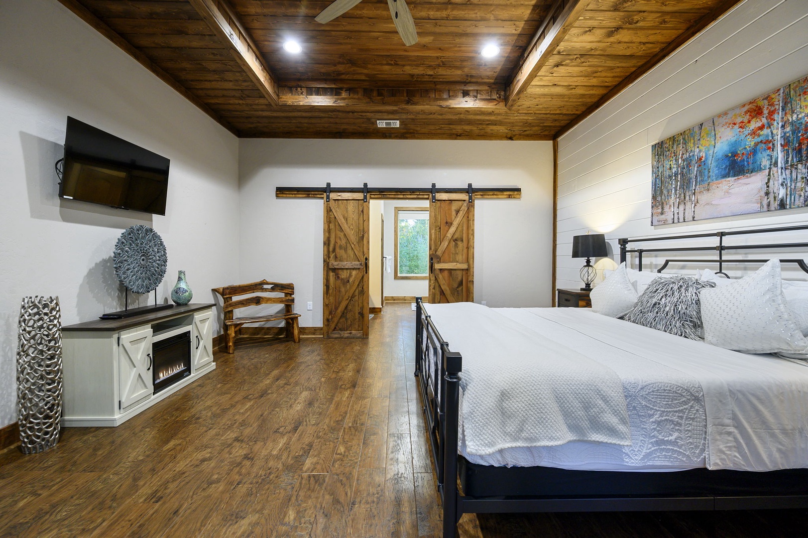 Your spacious bedroom features a plush bed, rustic barn doors, and elegant wood-beamed ceiling. Unwind with the TV and decorative fireplace, all crafted for your comfort.