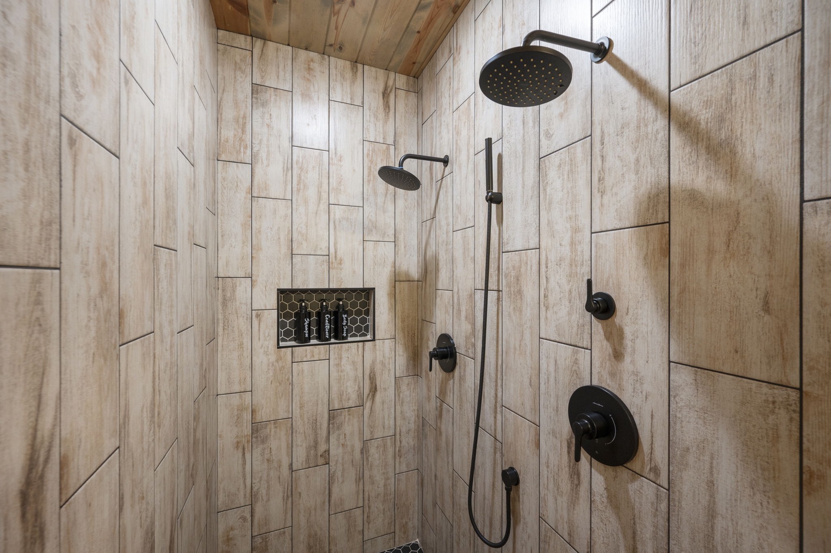 Your modern shower features double rainfall heads and a handheld option, perfectly framed by warm wood-look tile and natural ceiling beams that give this space a cozy yet contemporary feel.