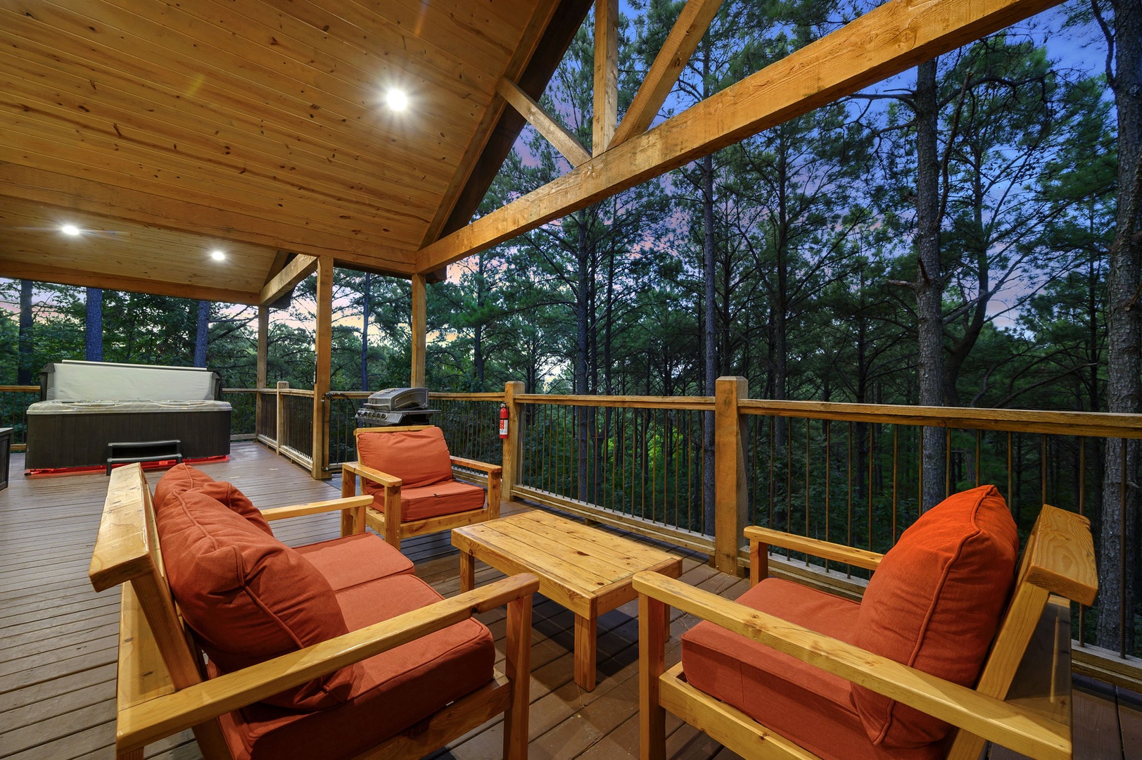 Your private covered deck overlooks lush forest, where warm lighting, cozy seating, and a hot tub create the perfect retreat for evening relaxation.