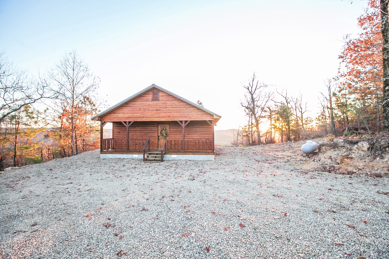Rustic wooden cabin nestled among autumn trees on a gravel clearing with mountain views in the distance.