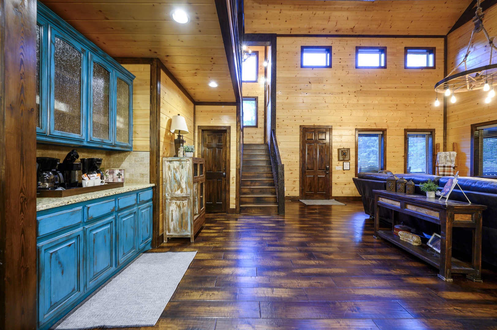 Open-concept retreat with rich wood floors, soaring ceilings, and charming turquoise kitchenette. Relax on the leather sofa as natural light floods through high windows, creating a warm, airy mountain escape.