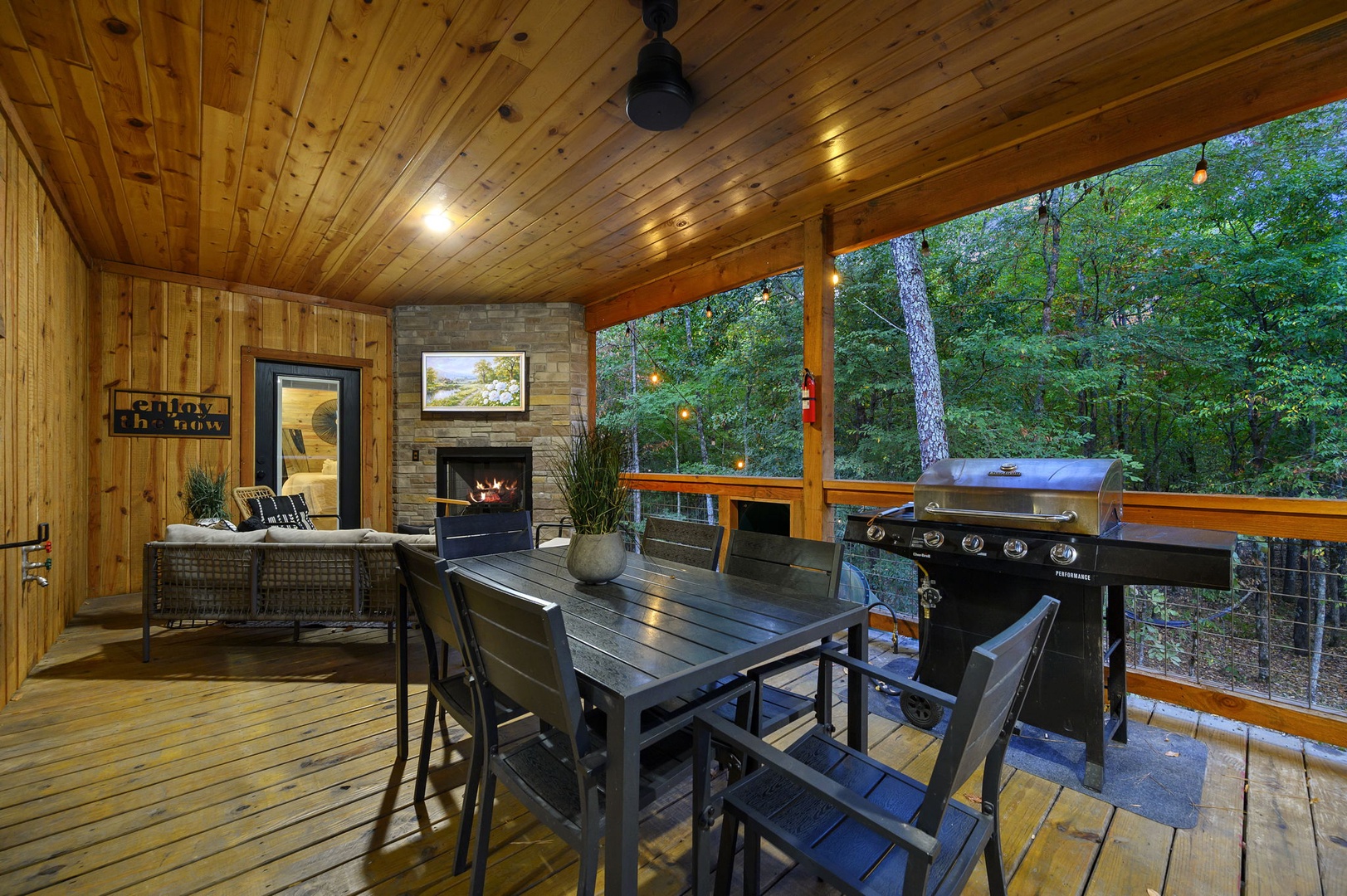 Your covered deck awaits with a crackling fireplace, dining space, and barbecue grill surrounded by peaceful forest views—perfect for evening gatherings under the warm glow of string lights.