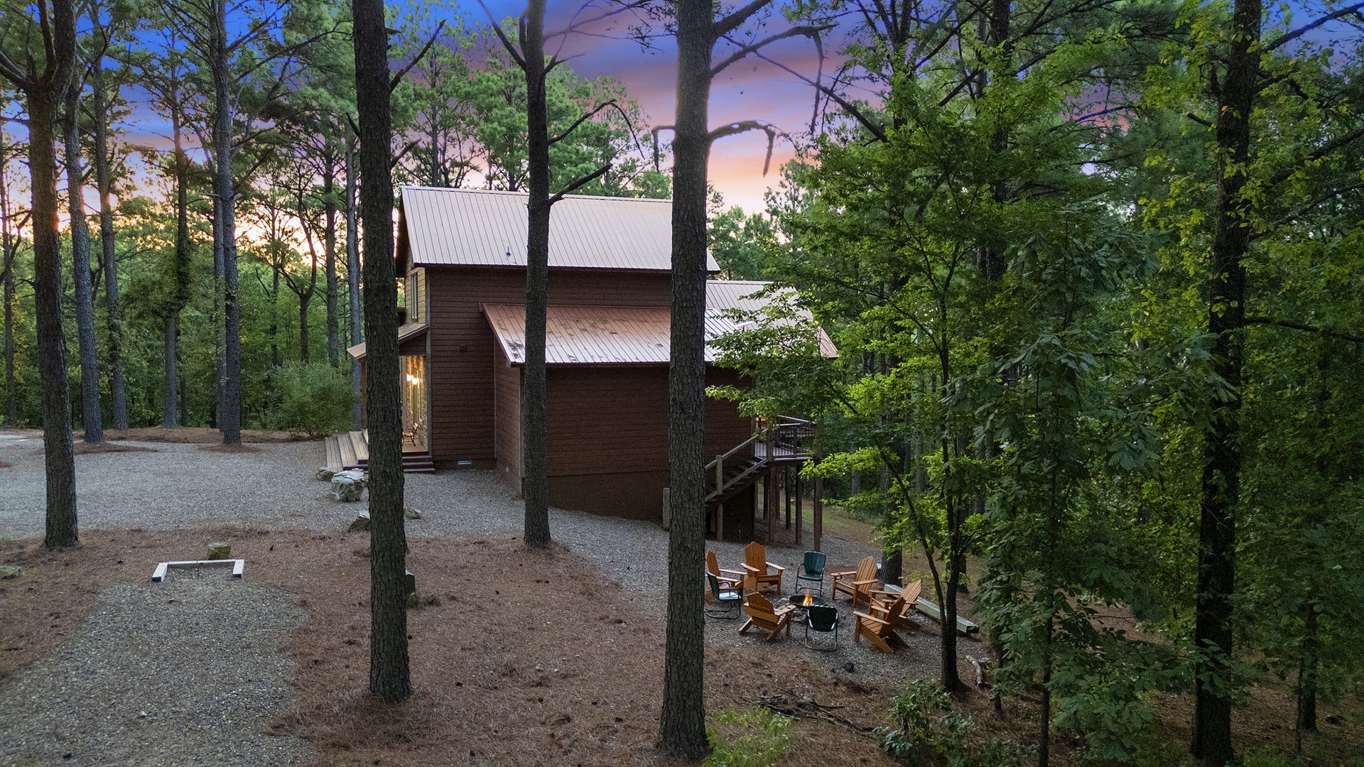 A secluded cabin nestled among tall trees with outdoor seating and a fire pit area, captured during a colorful sunset.