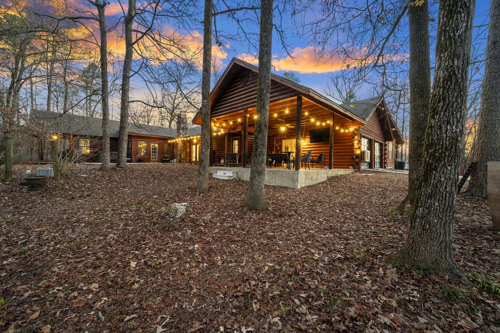 Rustic log cabin nestled in mature woodland with warm string lights creating magical evening ambiance against colorful twilight sky.