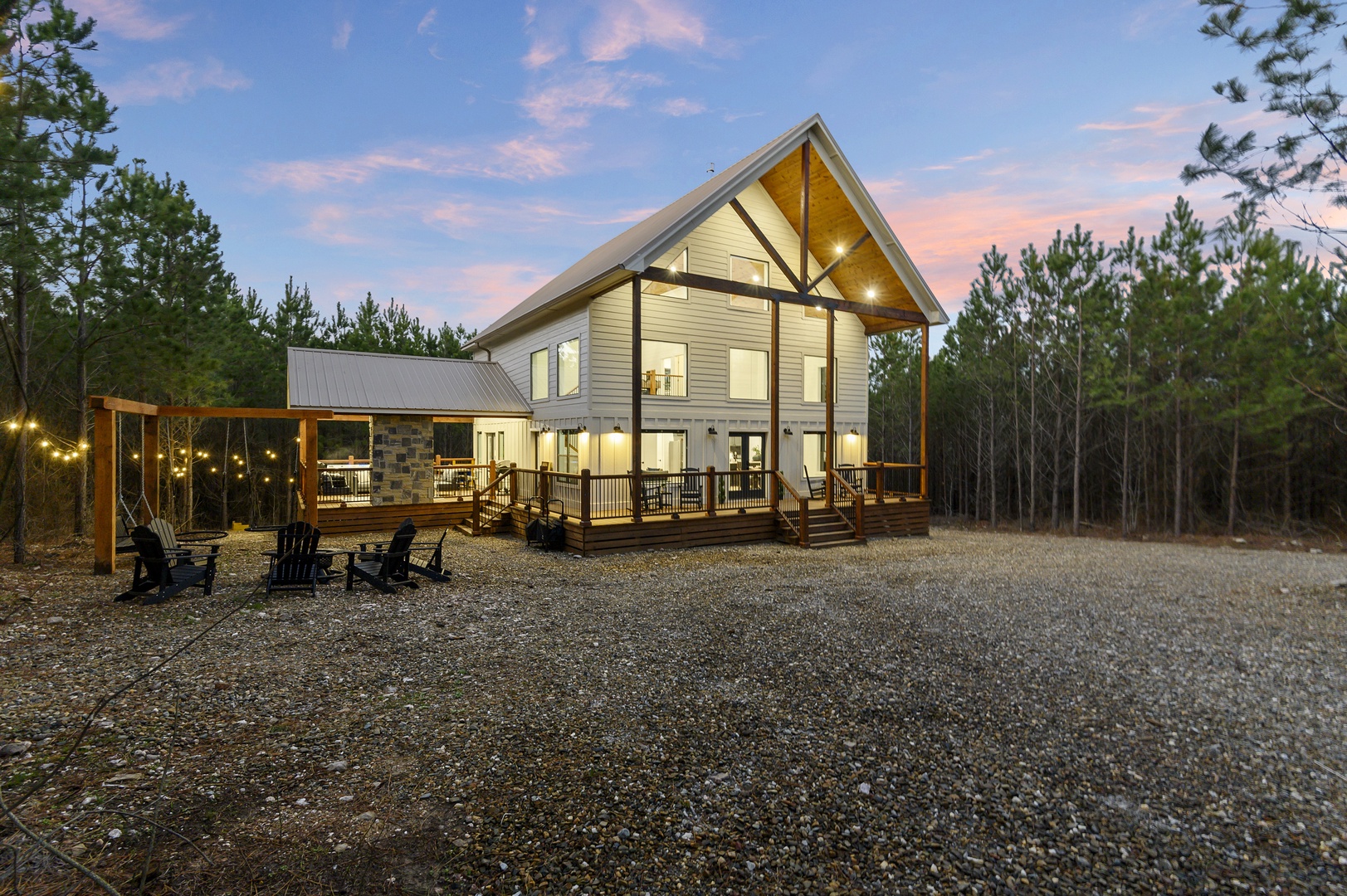 Modern mountain retreat with wraparound deck and fire pit area surrounded by towering pine forest at twilight.