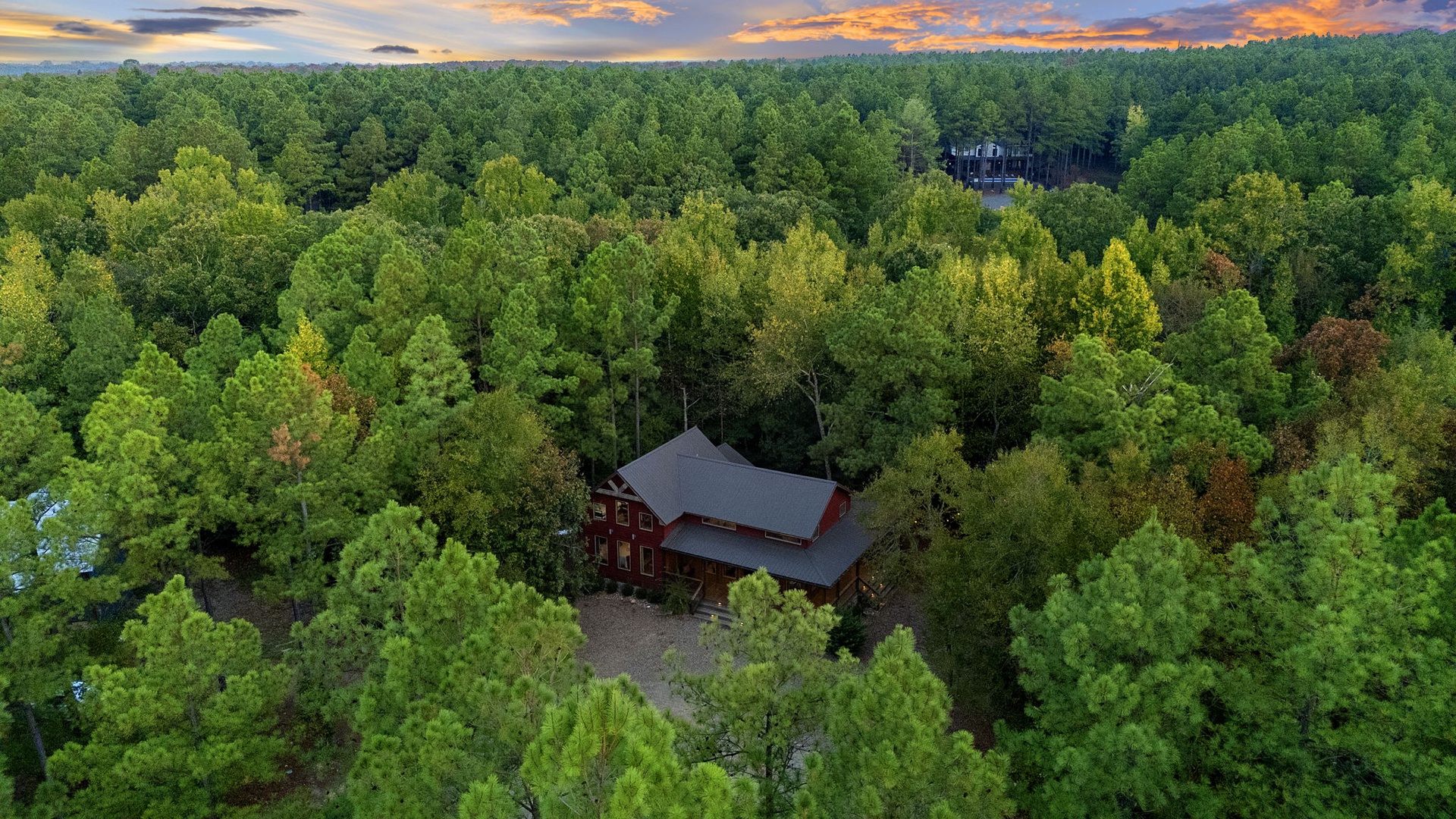 An aerial view of a secluded red lodge nestled among thick forest, with a dramatic sunset casting orange and purple hues across the sky above the trees.