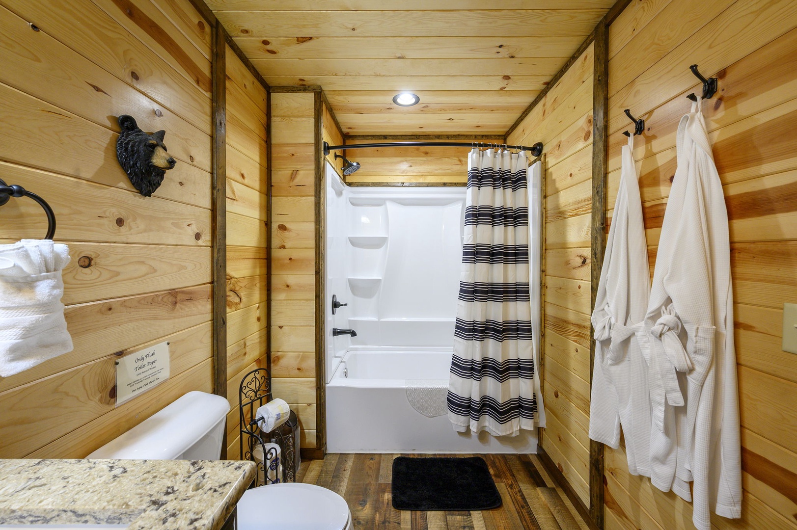 Wrap yourself in cozy cabin charm with this rustic bathroom featuring warm wood paneling, a refreshing shower-tub combo, and plush robes and towels ready for your use.